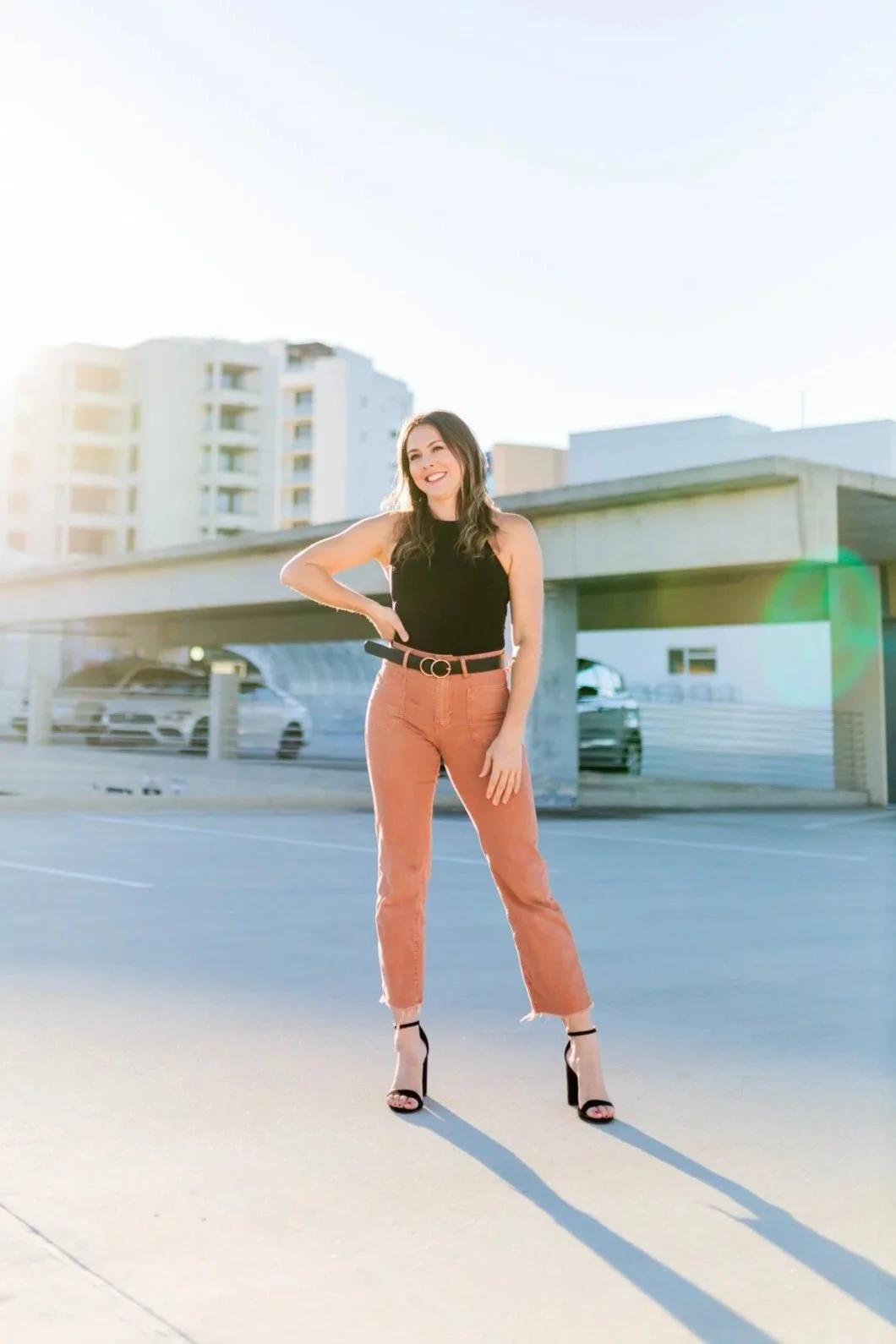 A woman, Lindsey Tosoni, standing in a parking lot during sunset, wearing a black sleeveless top, salmon-colored pants, and high heels, smiling and posing with her right hand on her hip.