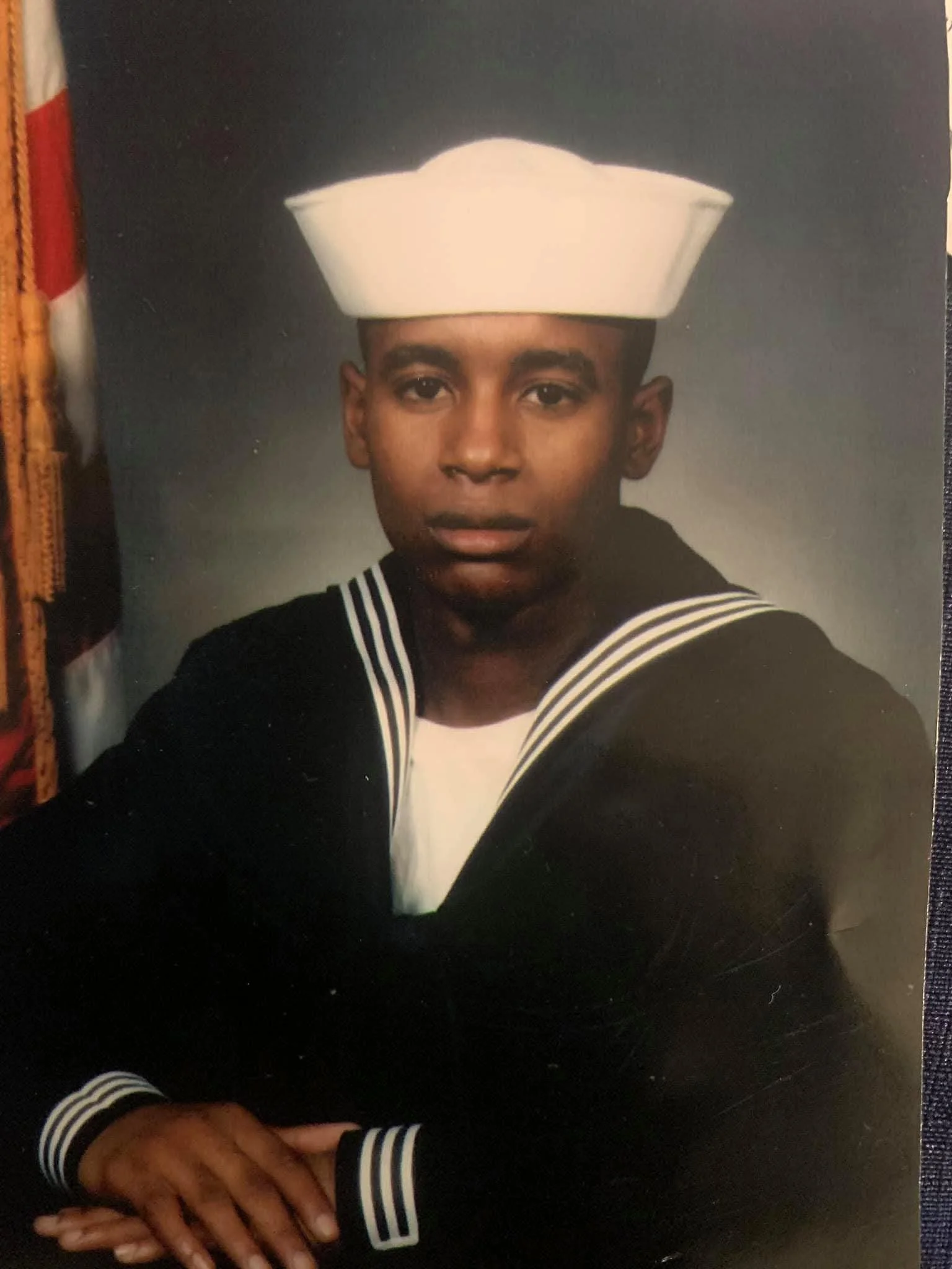 A young man in a United States Navy dress uniform, wearing a white sailor cap, posing with his hands folded.