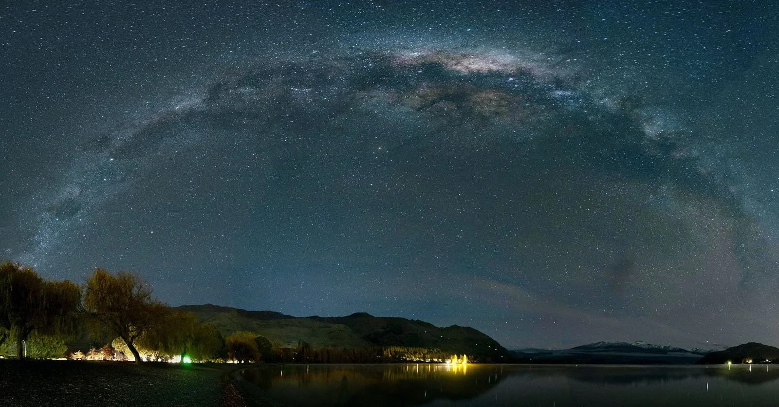 Night sky filled with stars and the Milky Way galaxy over a lake, with forested hills and mountains in the background, and a few small light sources on the shoreline.