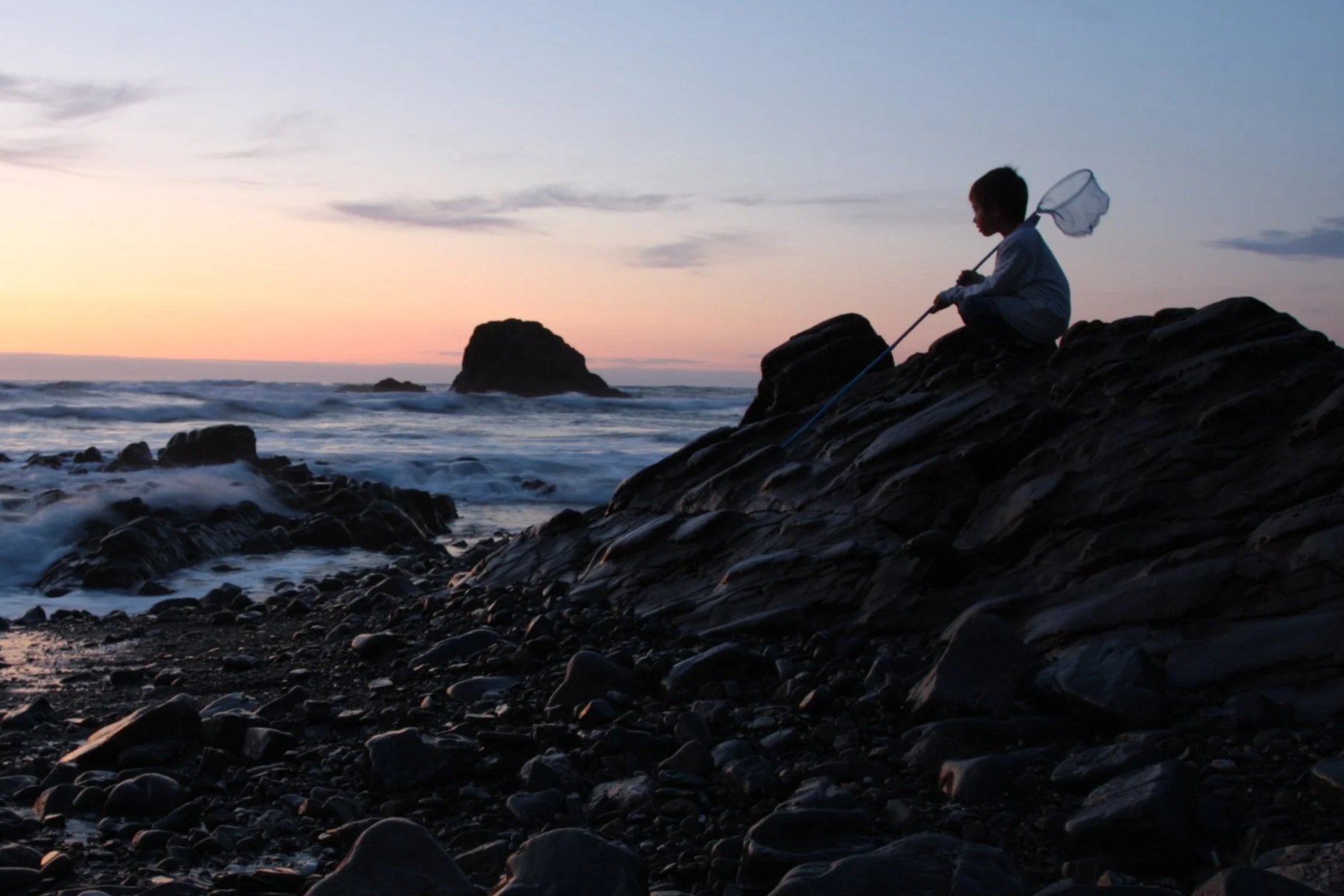 A child sitting on rocks along the shoreline at sunset, holding a net, with waves crashing nearby and a large rock formation in the background.