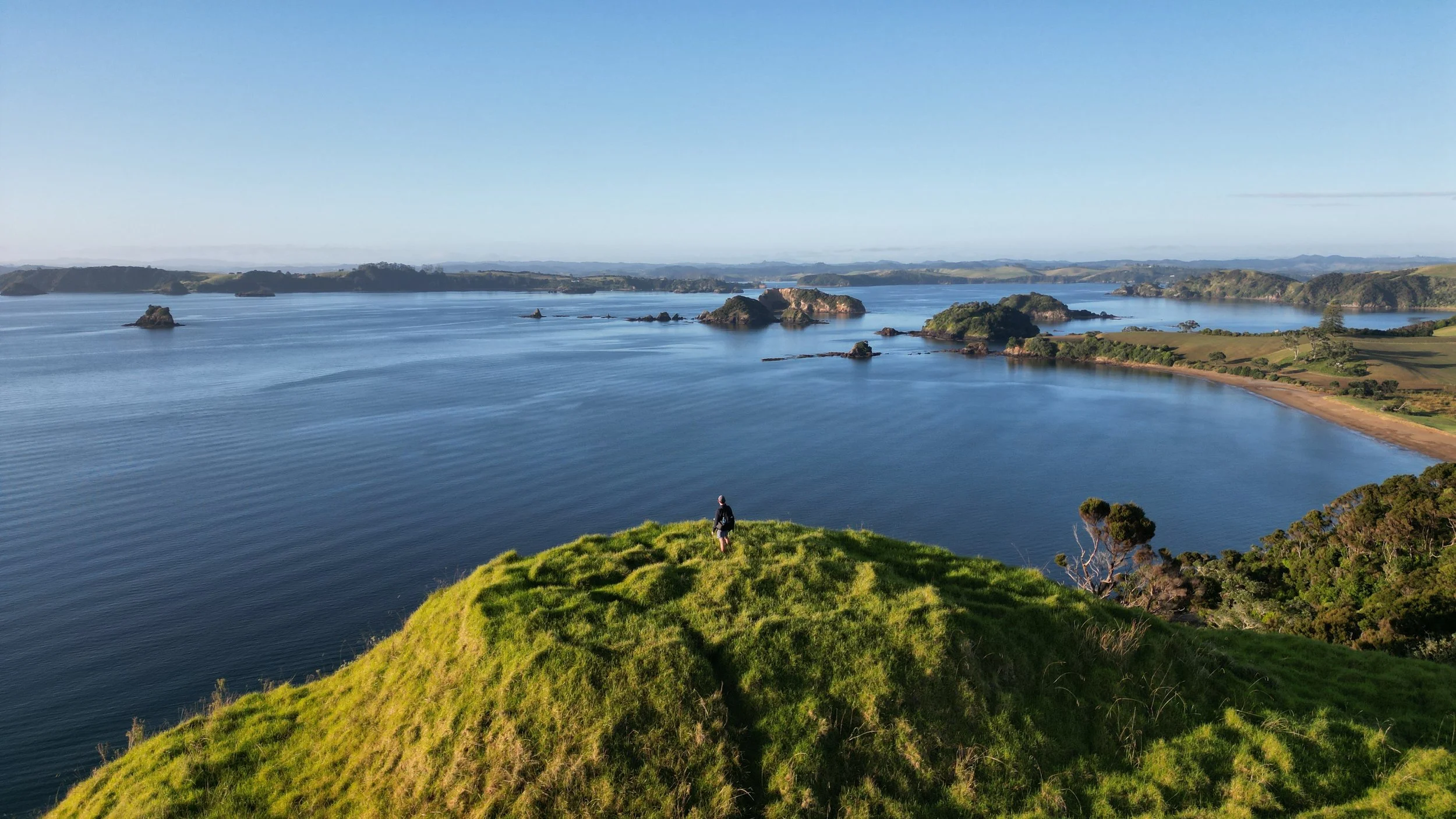 A person standing on a grassy hill overlooking a large body of water with numerous small islands and a distant shoreline.