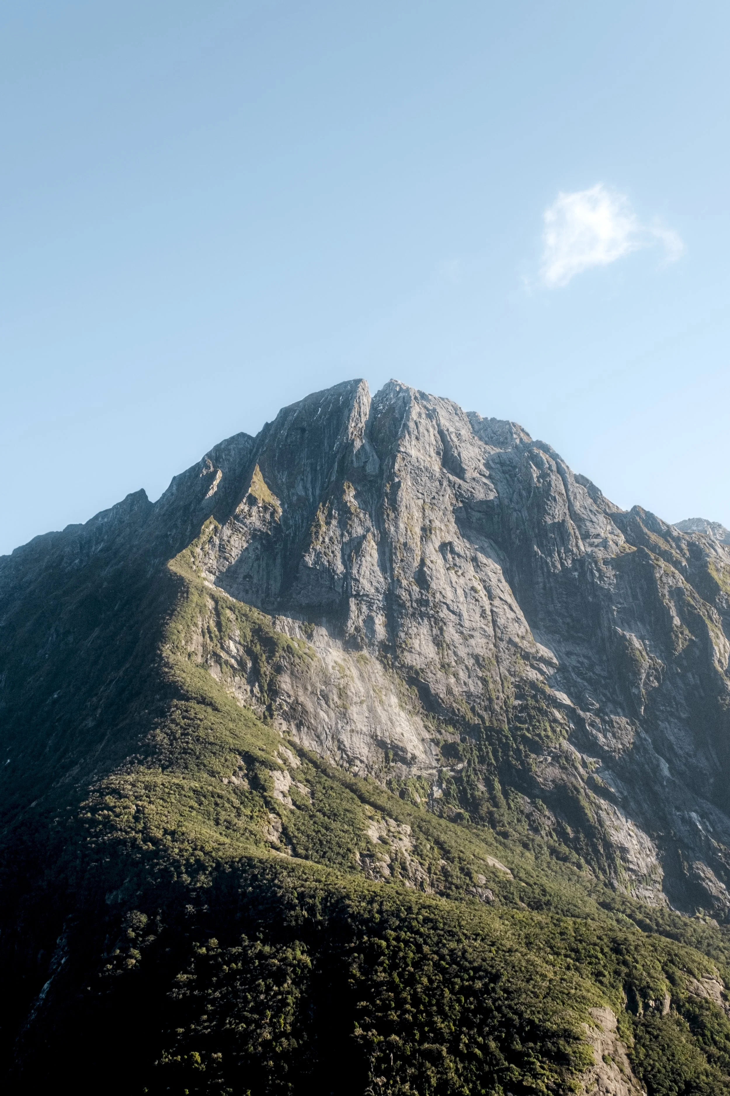 Mountain with rocky peaks and green vegetation on the slopes, under a clear blue sky with a single small cloud.