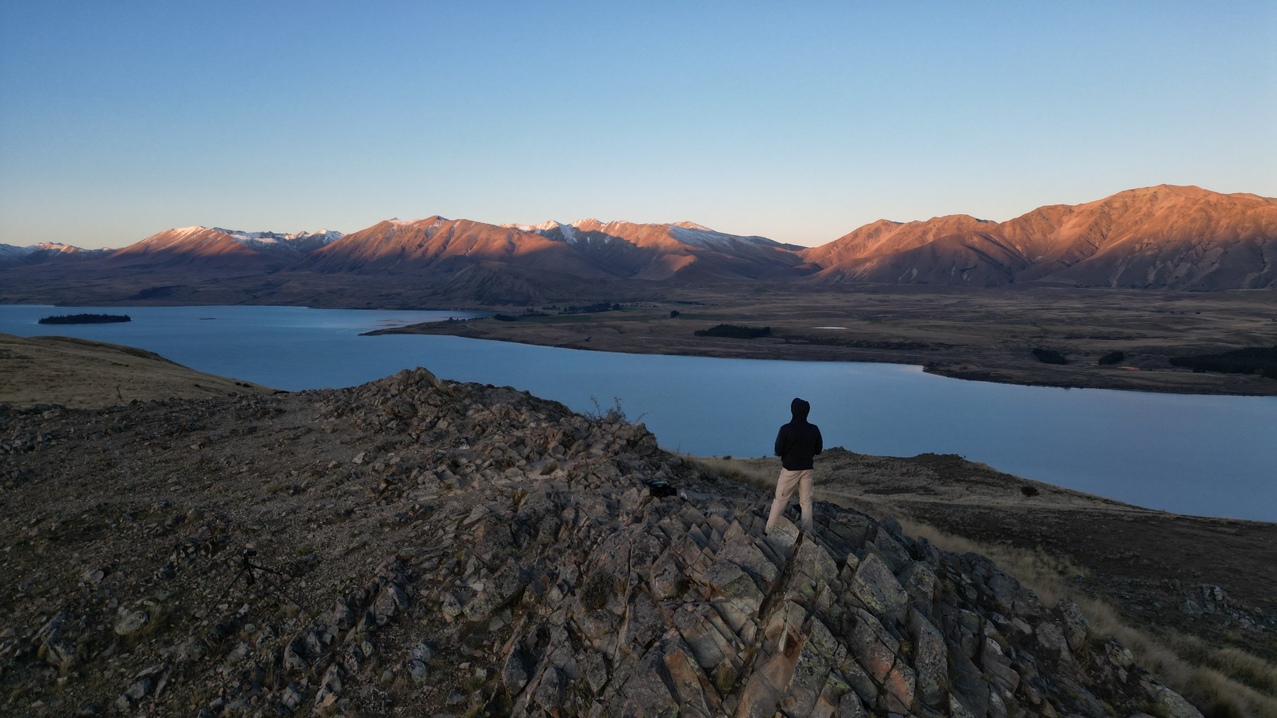 A person standing on rocky terrain overlooking a large river with mountains in the background during sunset.