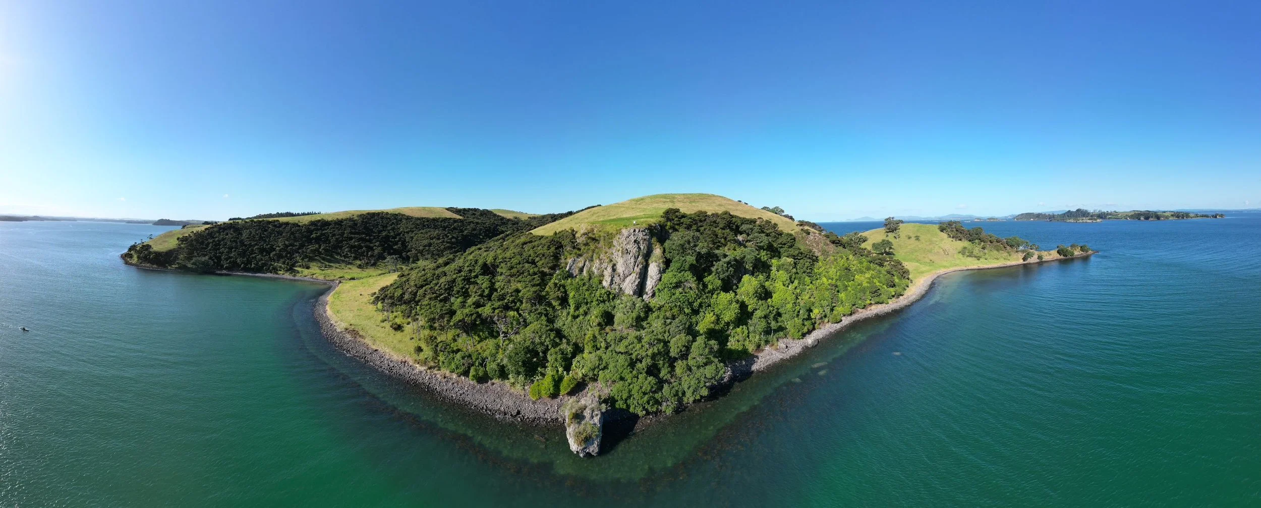 Aerial view of a lush, green island surrounded by blue water under clear sky, with rolling hills and rocky coastlines.