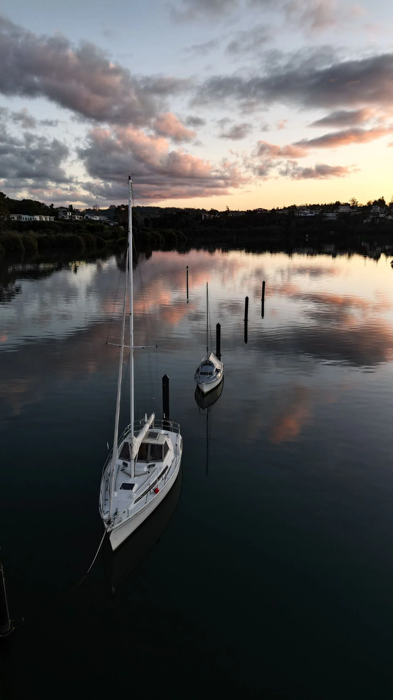 Sailboats docked on calm water during sunset with cloudy sky and houses on the hillside in the background.