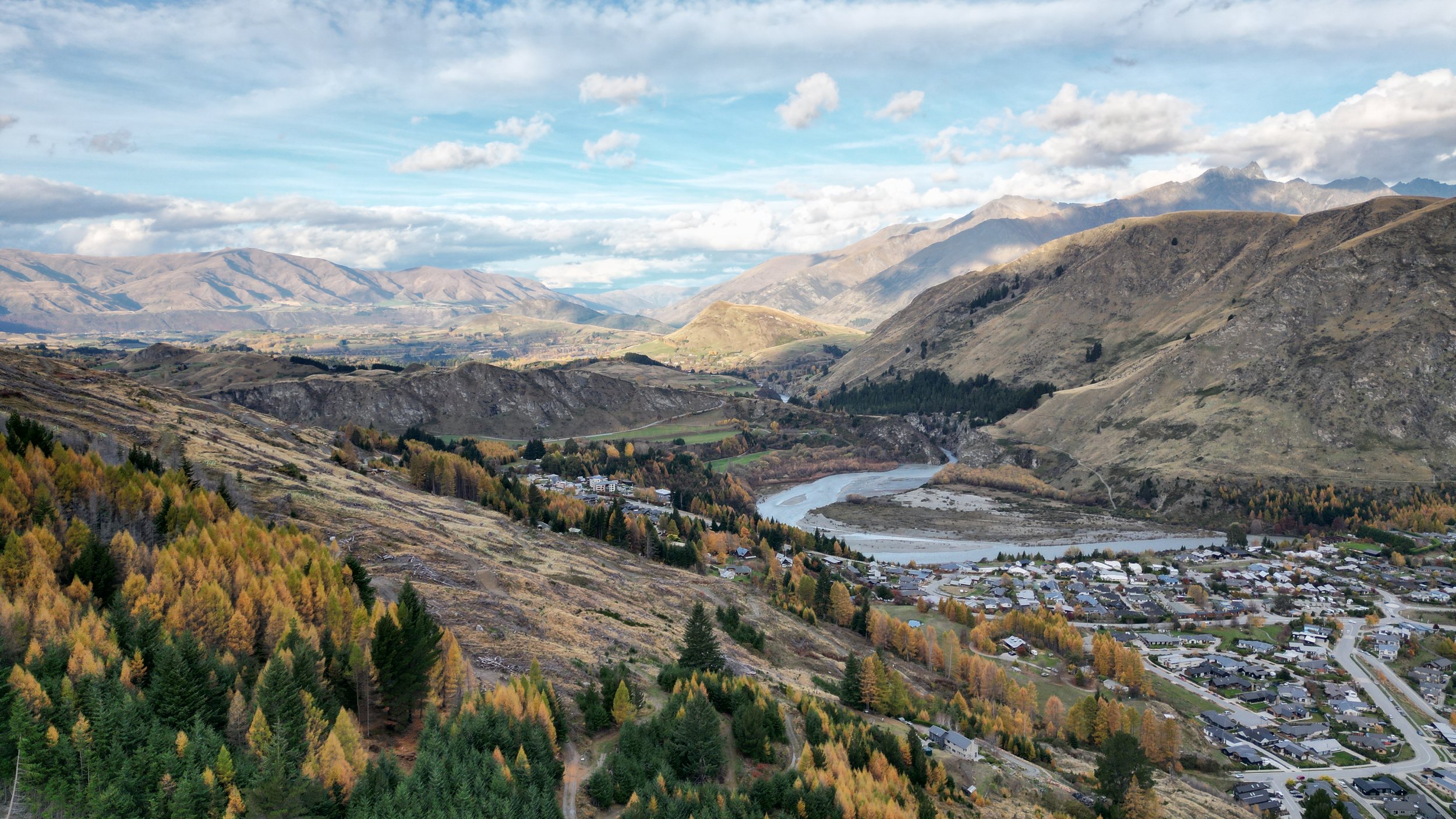 A scenic landscape of rolling mountains and valleys with a river running through a small town, under a partly cloudy sky.