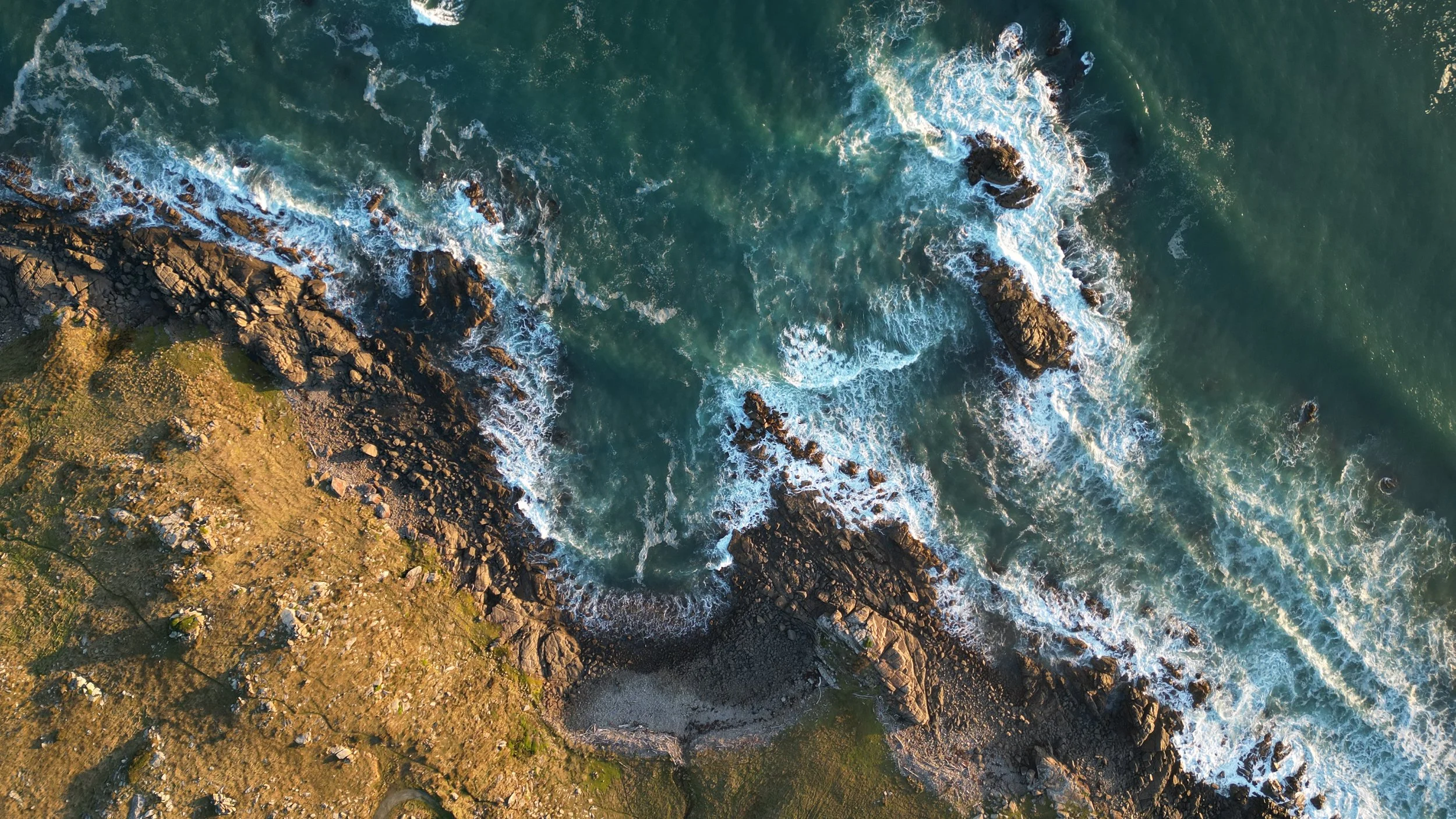 Aerial view of rocky coastline with waves crashing onto the shore, with green grassy areas and rocks visible.