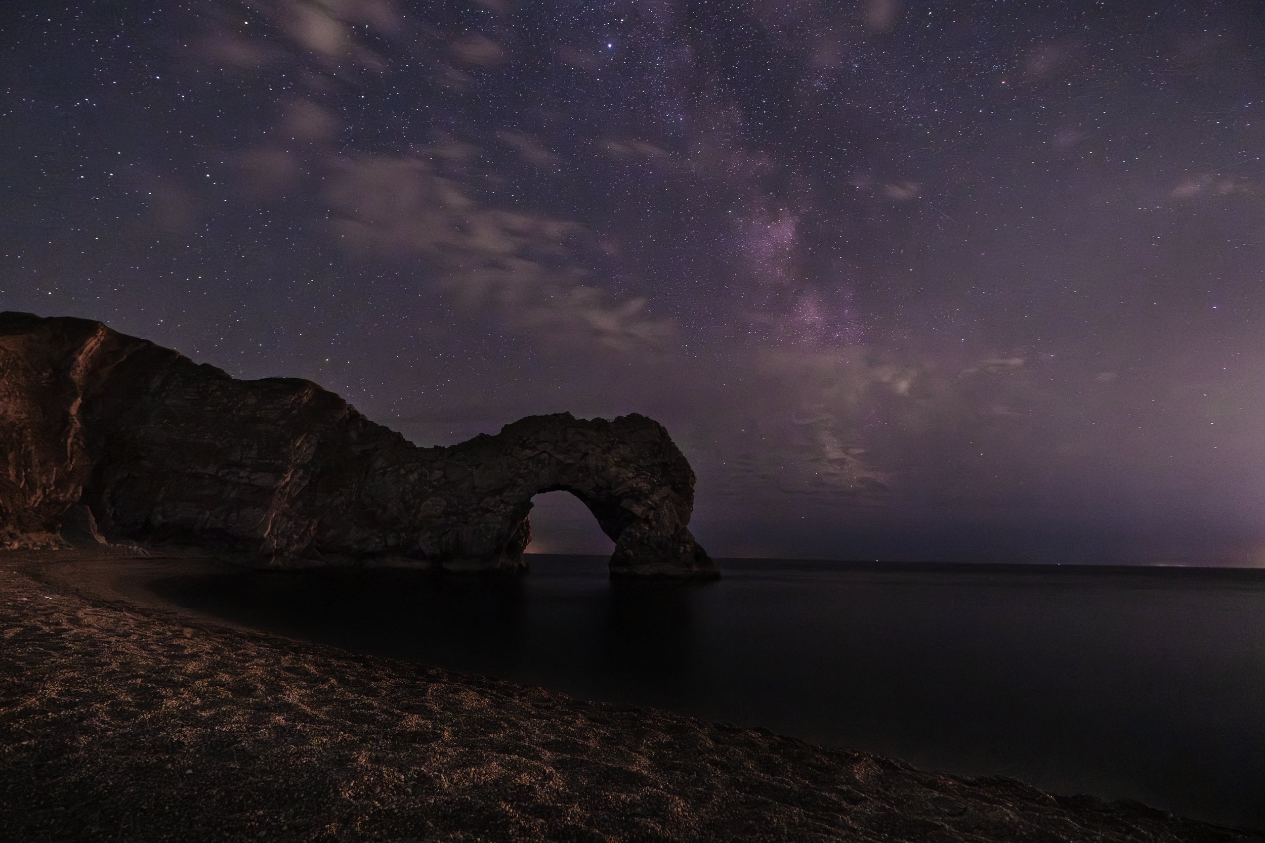 Nighttime view of a starry sky above a rocky beach with a natural rock arch formation.