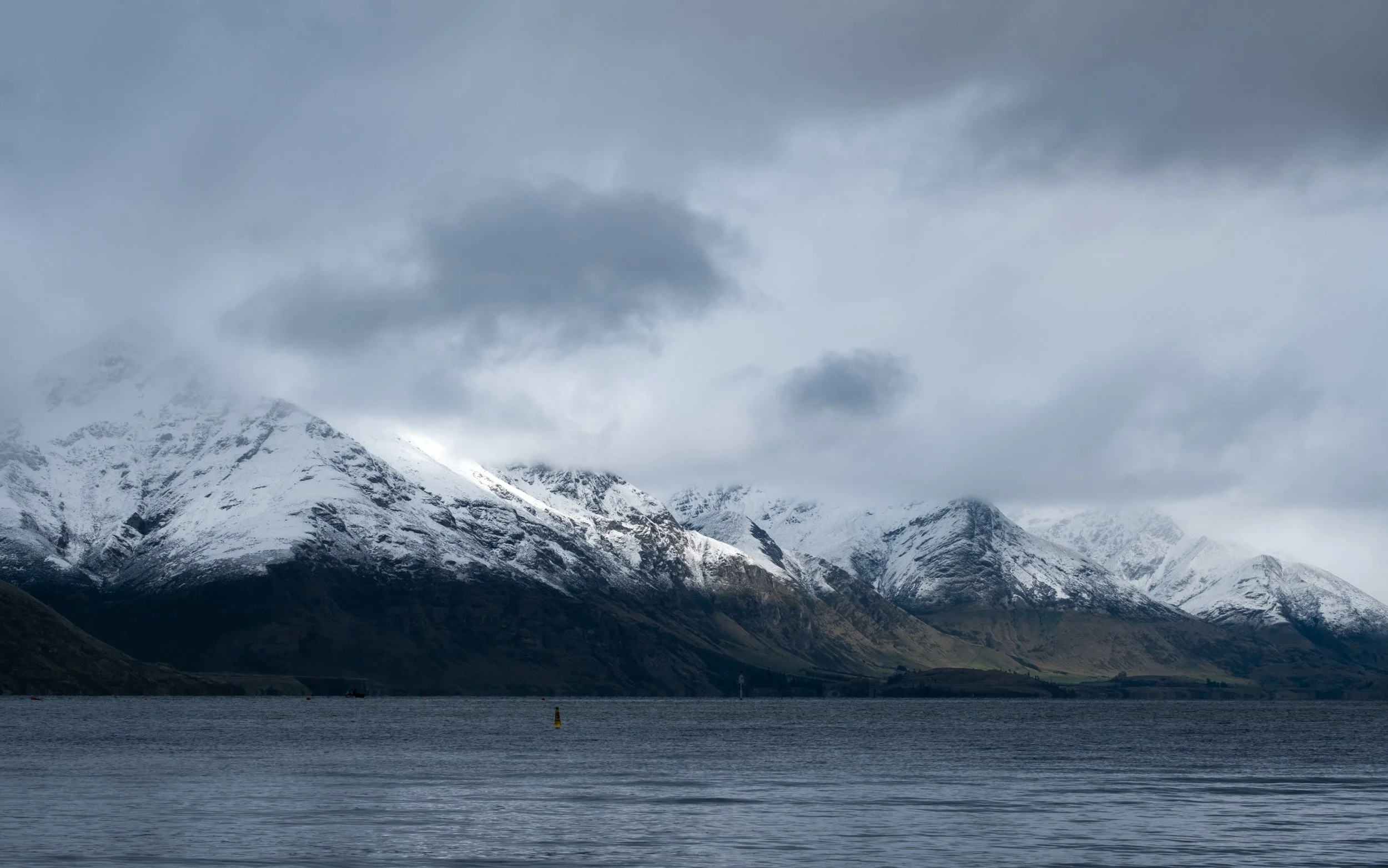 Snow-capped mountains partially covered in clouds seen across a calm body of water with a few buoys floating on the surface.