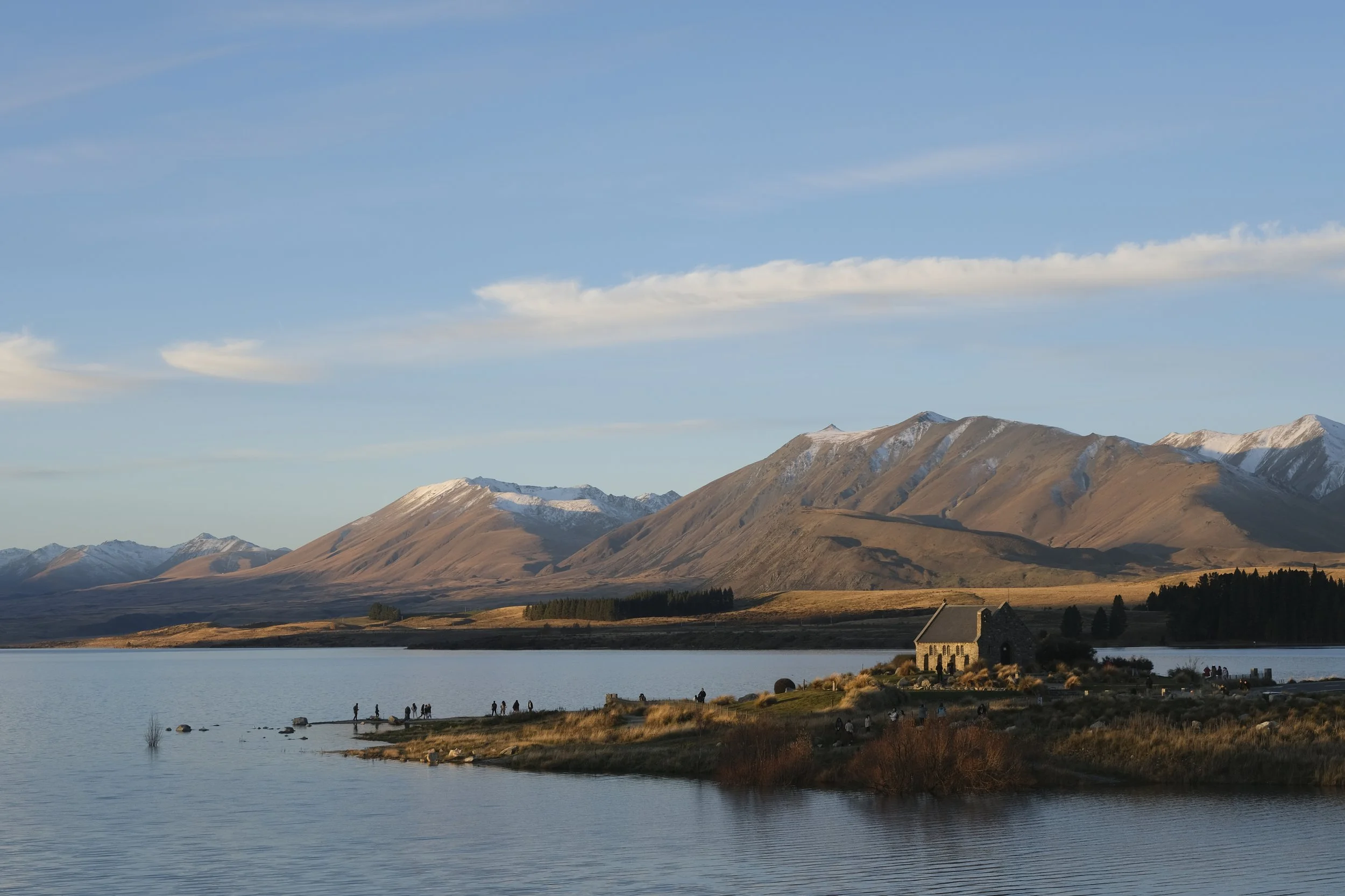 Scenic landscape with a lake in the foreground, a small stone house on the shore, mountains in the background, and a blue sky with clouds overhead.