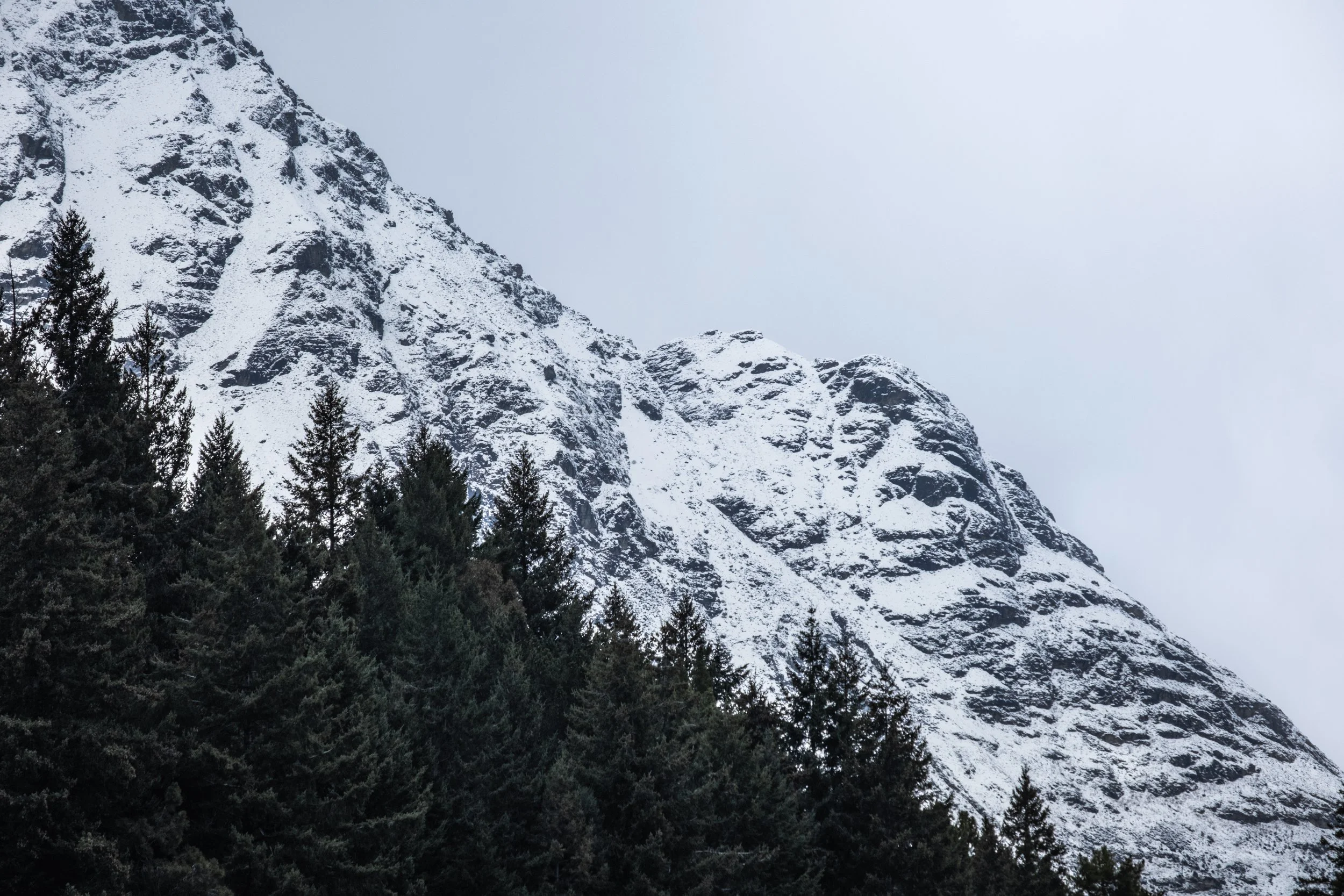 Snow-covered mountain with dark green trees in the foreground under a gray cloudy sky.