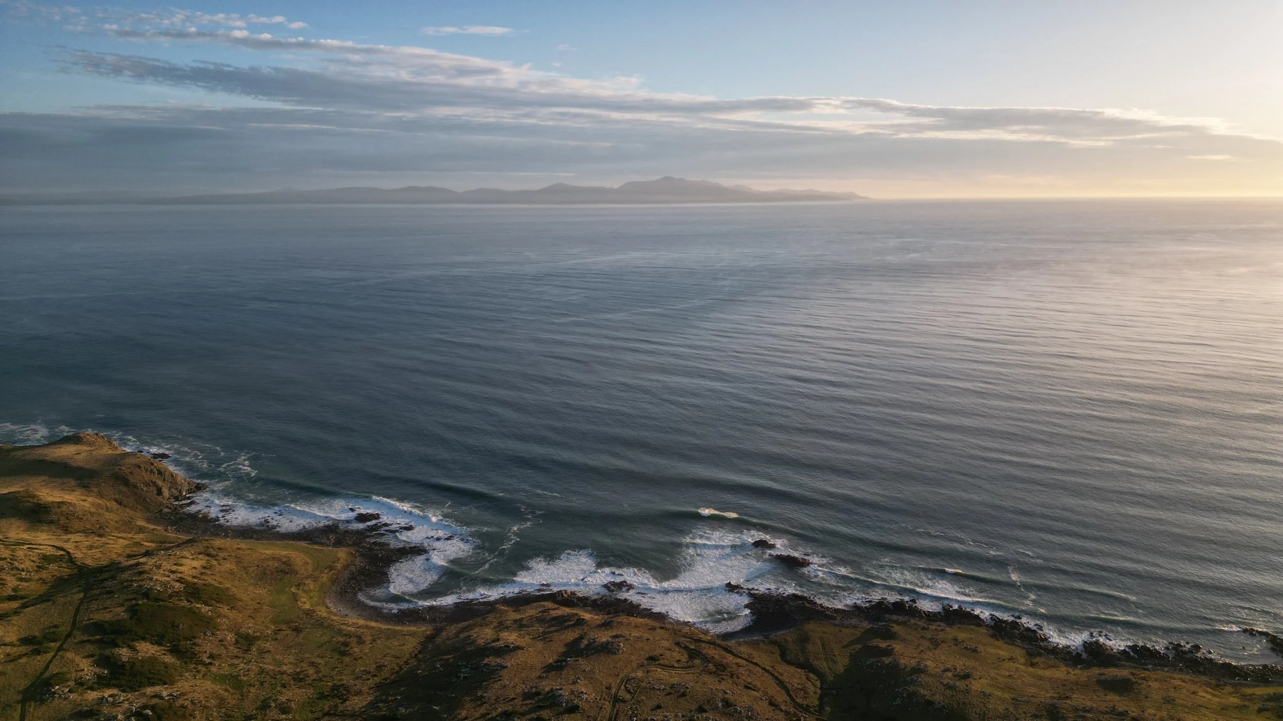A scenic view of the ocean with a distant landmass on the horizon, cloudy sky, and a rugged coastline in the foreground.