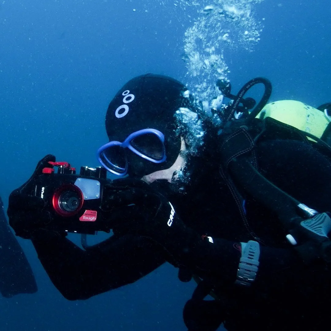 A scuba diver underwater holding an underwater camera, wearing a mask, snorkel, and black wetsuit.
