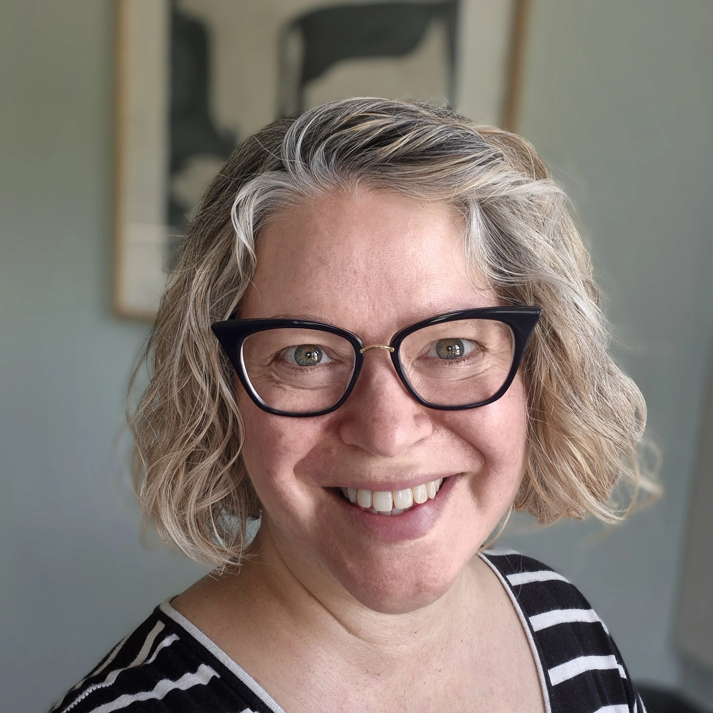A portrait photo of a light skinned woman with wavy short blonde hair, smiling, wearing dark framed glasses and a black and white striped top