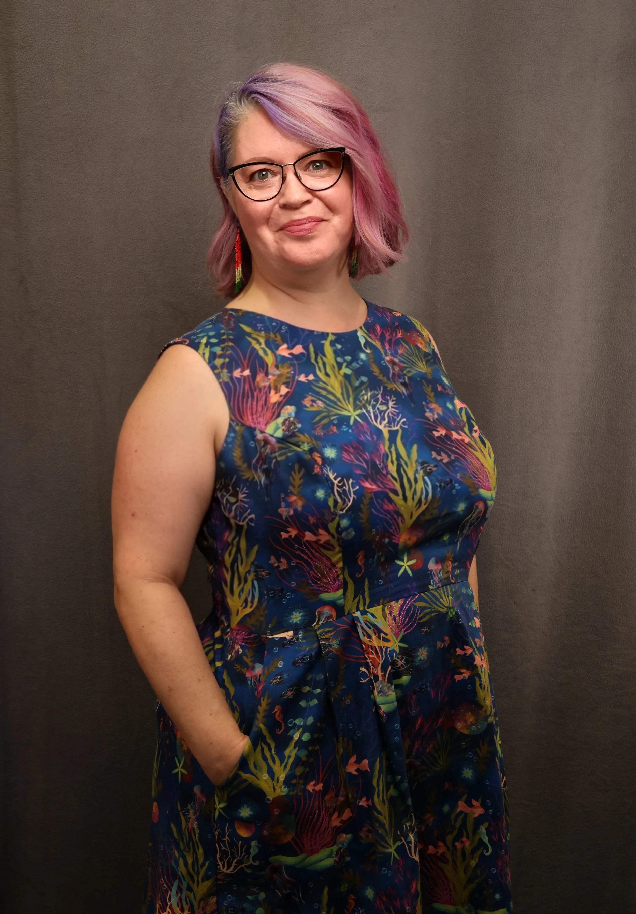 Professional portrait of a light-skinned woman with rose gold short hair and dark-rimmed glasses. She is wearing a sleeveless colourful dress and has her hands in her pockets and a smile on her face.