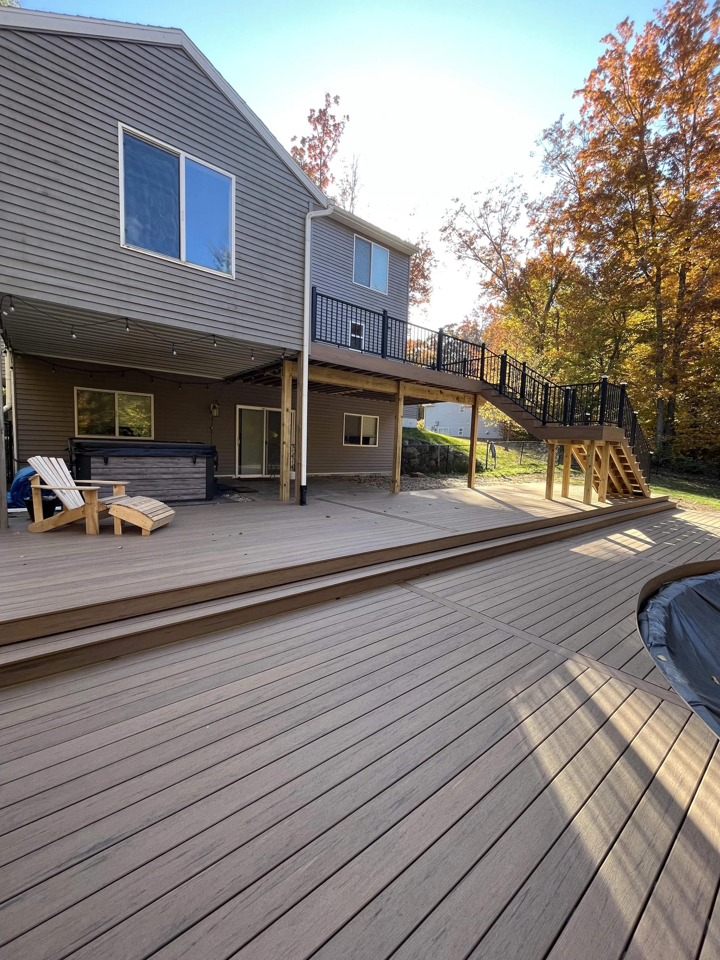 Backyard with a large wooden deck, outdoor furniture, staircase, and a house with gray siding, surrounded by trees with autumn leaves.