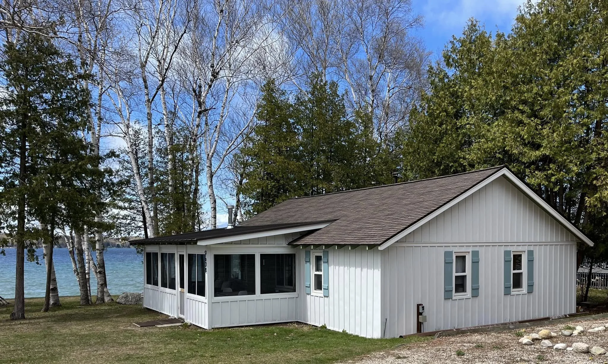A white house with blue shutters and a brown roof located near a lake, surrounded by tall trees, some with green leaves and others with bare branches, under a partly cloudy blue sky.