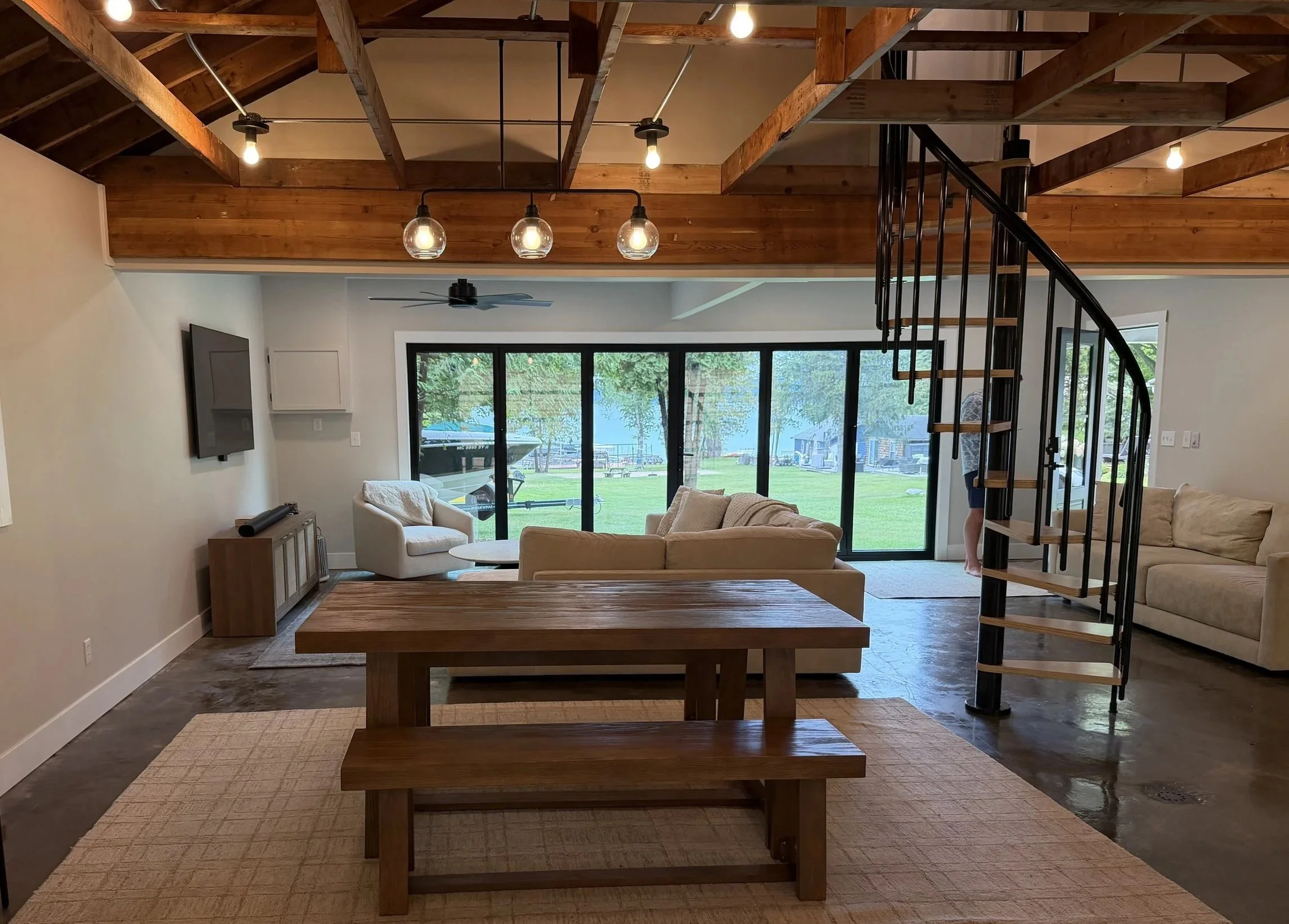 Living room with wooden staircase, ceiling lights, ceiling fan, sliding glass doors leading outside, beige sofas, and a wooden dining table.