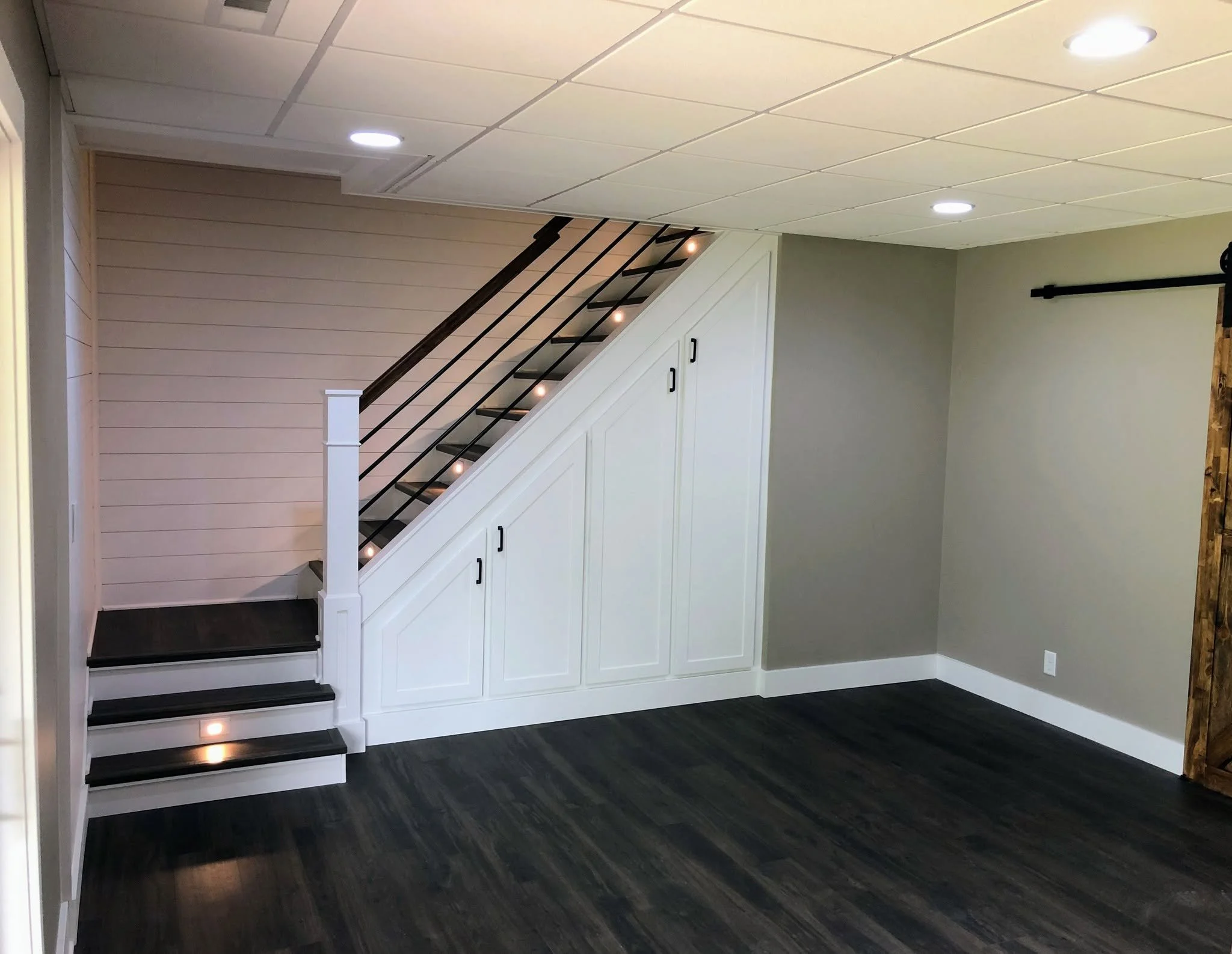 Interior of a modern basement or living space featuring a staircase with black metal railings and white risers, built-in storage cabinets underneath the stairs, a white paneled wall, gray walls, wooden accents, and recessed ceiling lights.