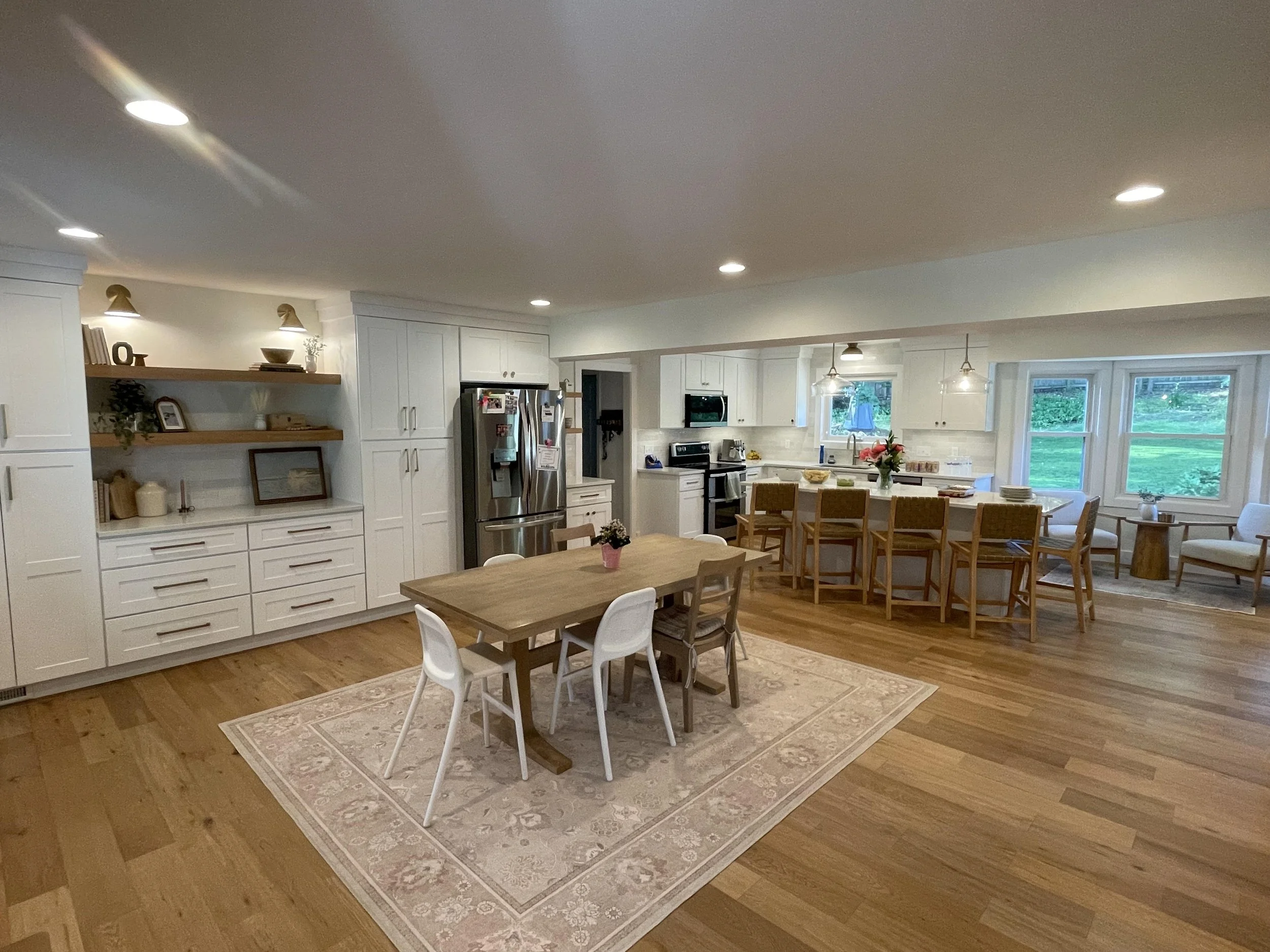 Bright, modern open-concept kitchen and dining area with white cabinets, stainless steel appliances, wooden dining table, chairs, and large windows showing a green backyard.