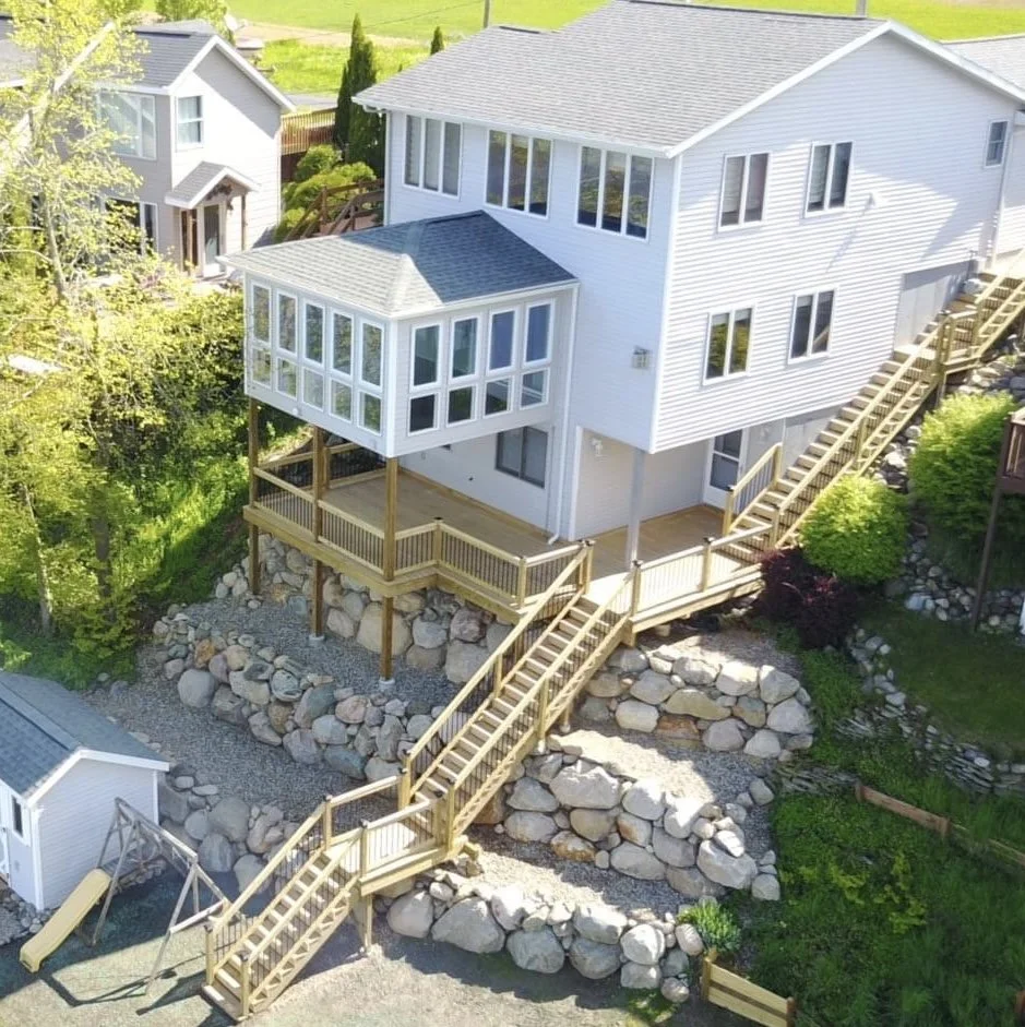 A white multi-story house with large windows and a wooden deck overlooking a landscaped yard with rocks and stairs.
