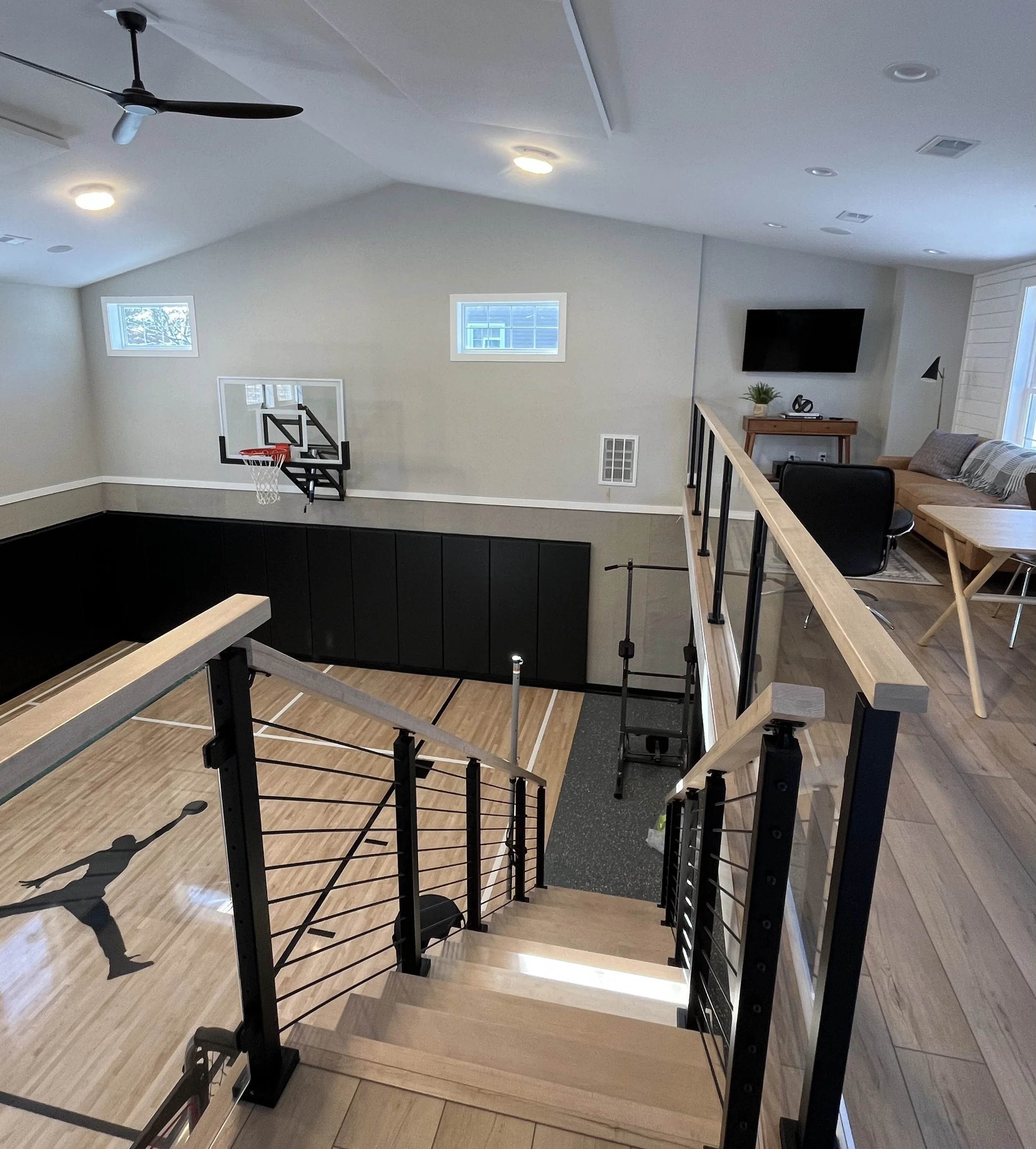 A staircase with a wooden handrail and black metal supports, leading down to a lower level with a basketball hoop mounted on a light gray wall, and some small windows near the ceiling. To the right, part of a living room area with a sofa, a black desk chair, a small wooden table, and a wall-mounted TV.