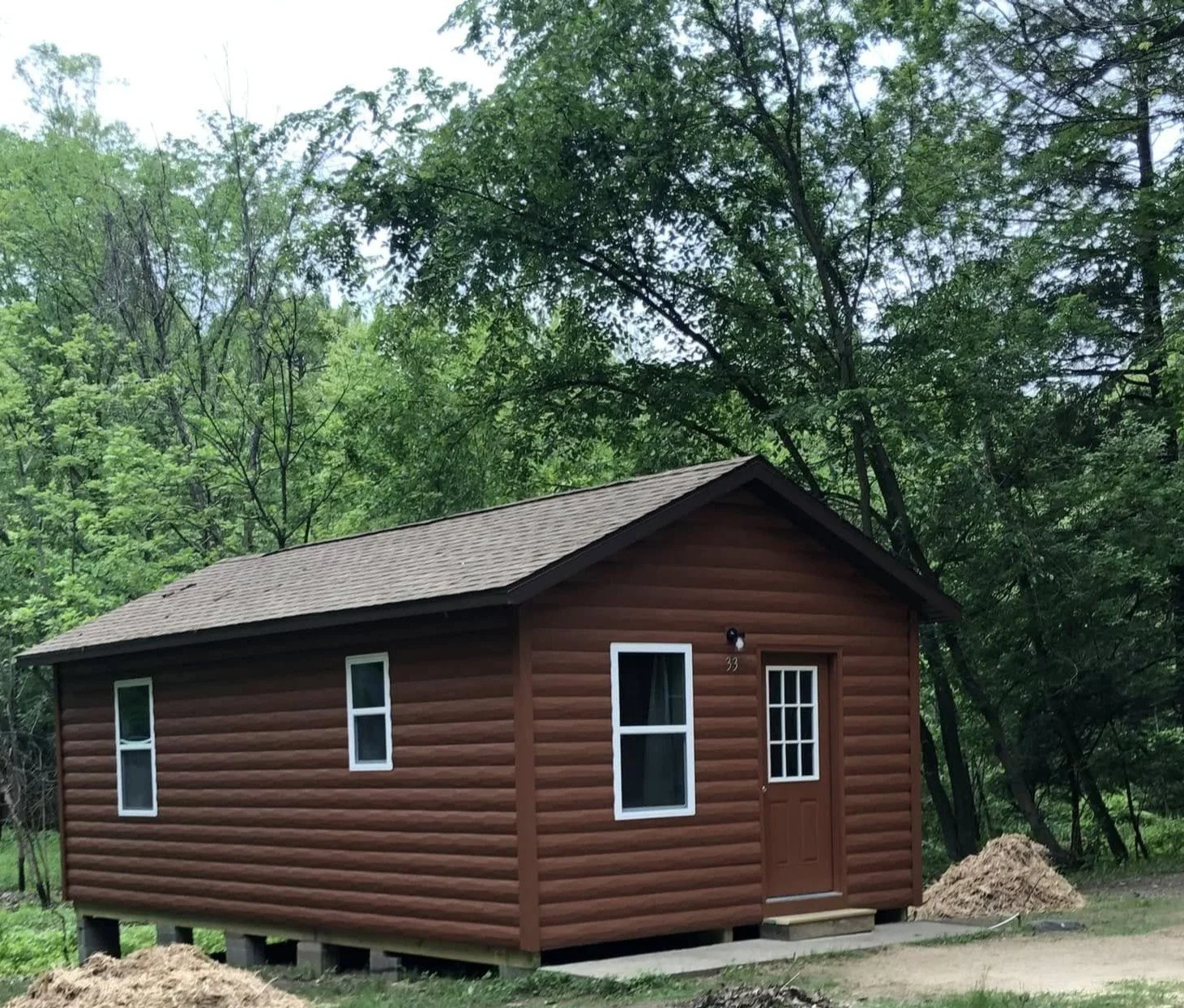 Small brown cabin with white-framed windows and door, surrounded by green trees, on a grassy area.