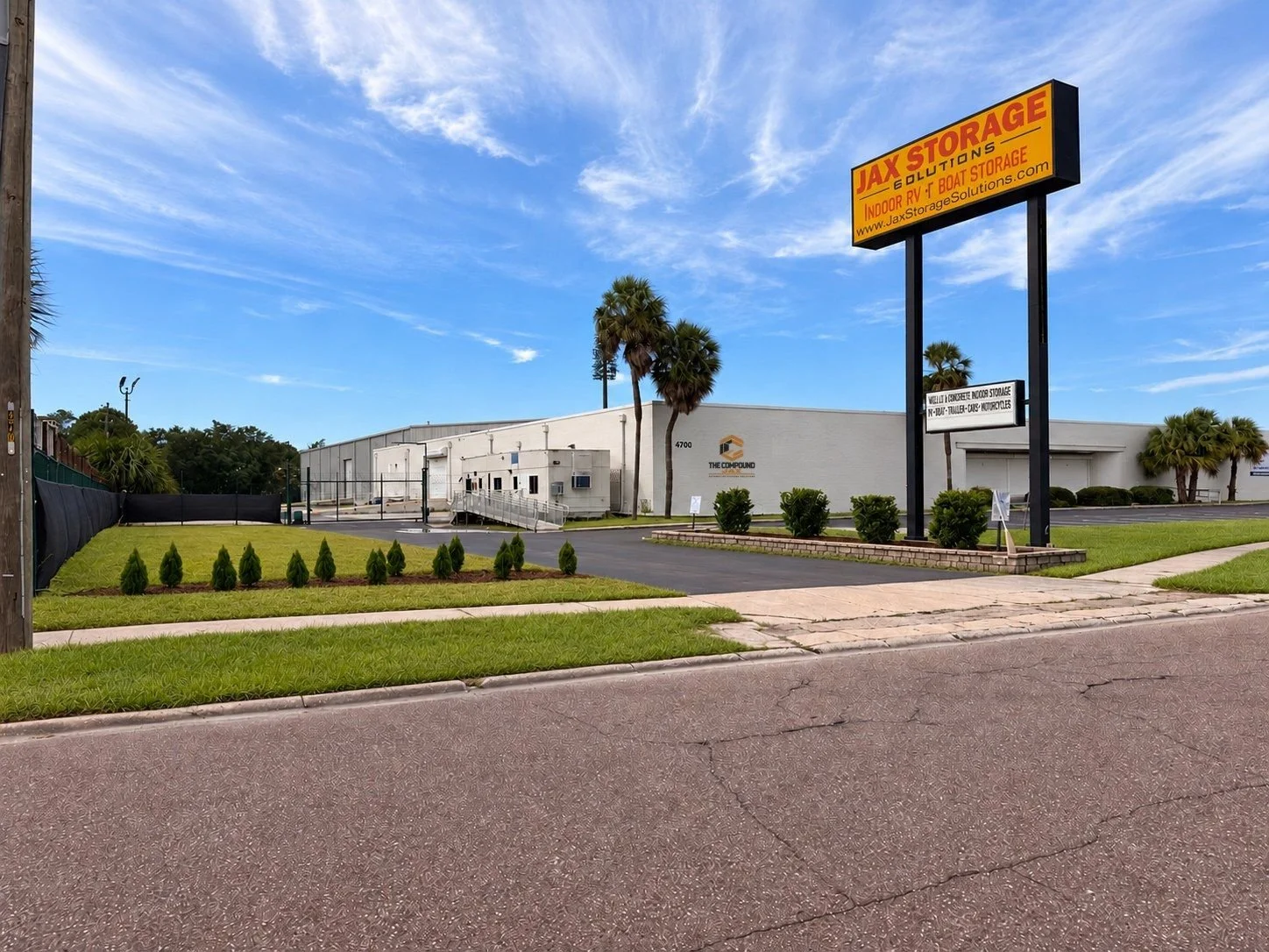 Exterior of a storage facility with a large yellow sign that reads 'JAX STORAGE & CONTAINERS, Indoor RV, Boat Storage'. The building is white with a landscaped area in front, including small bushes and young trees, under a blue sky with some clouds.