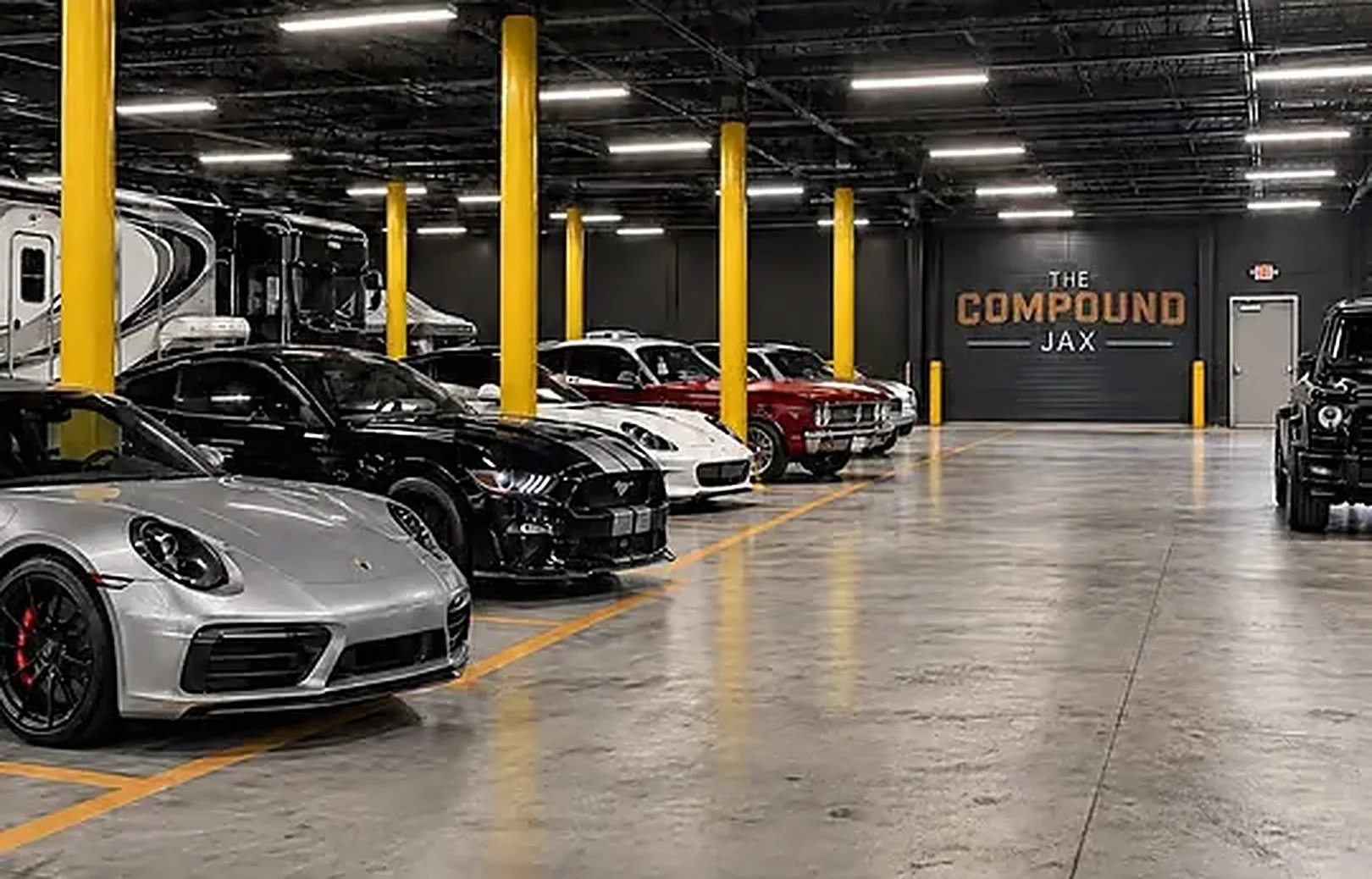 Indoor parking garage with a row of luxury cars including a silver Porsche, black Ford Mustang, white and red classic cars, and other vehicles, with yellow pillars and a sign on the back wall that reads 'The Compound JAX'.