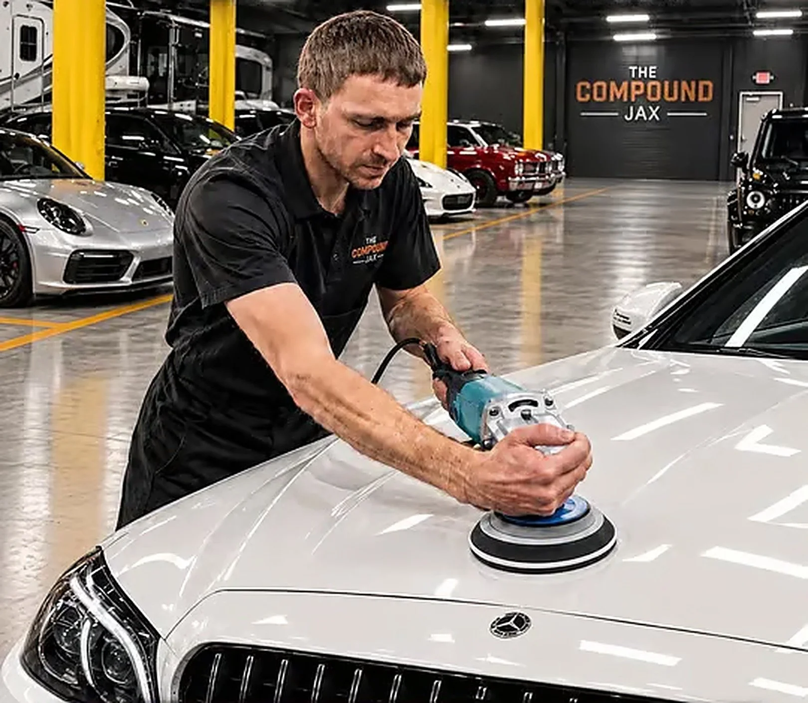 A man polishing the hood of a white Mercedes-Benz inside a car dealership or showroom with multiple luxury cars parked in the background and a sign that reads 'The Compound Jax'.