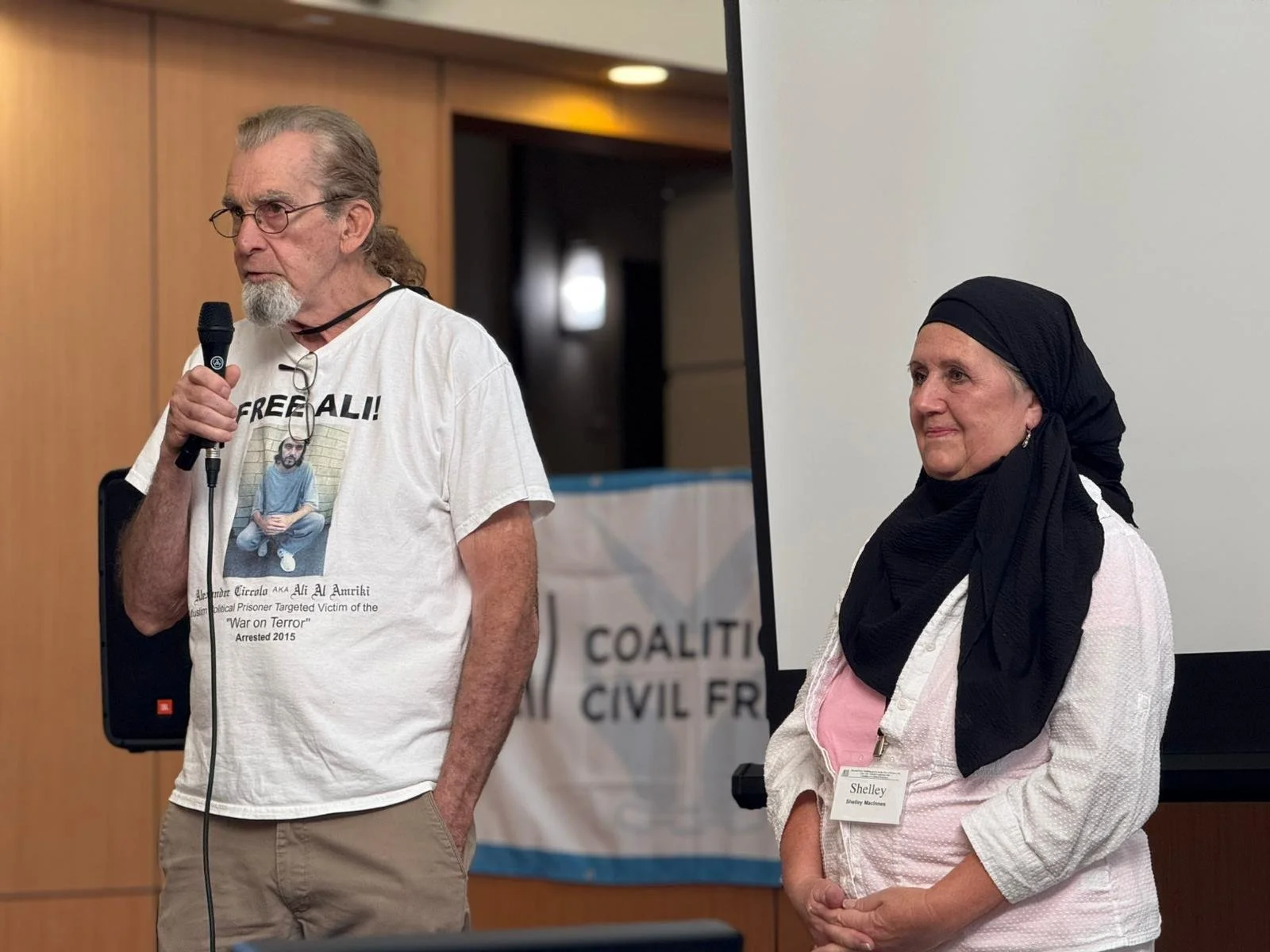 Man speaking into a microphone beside a woman wearing a black headscarf at a public presentation.