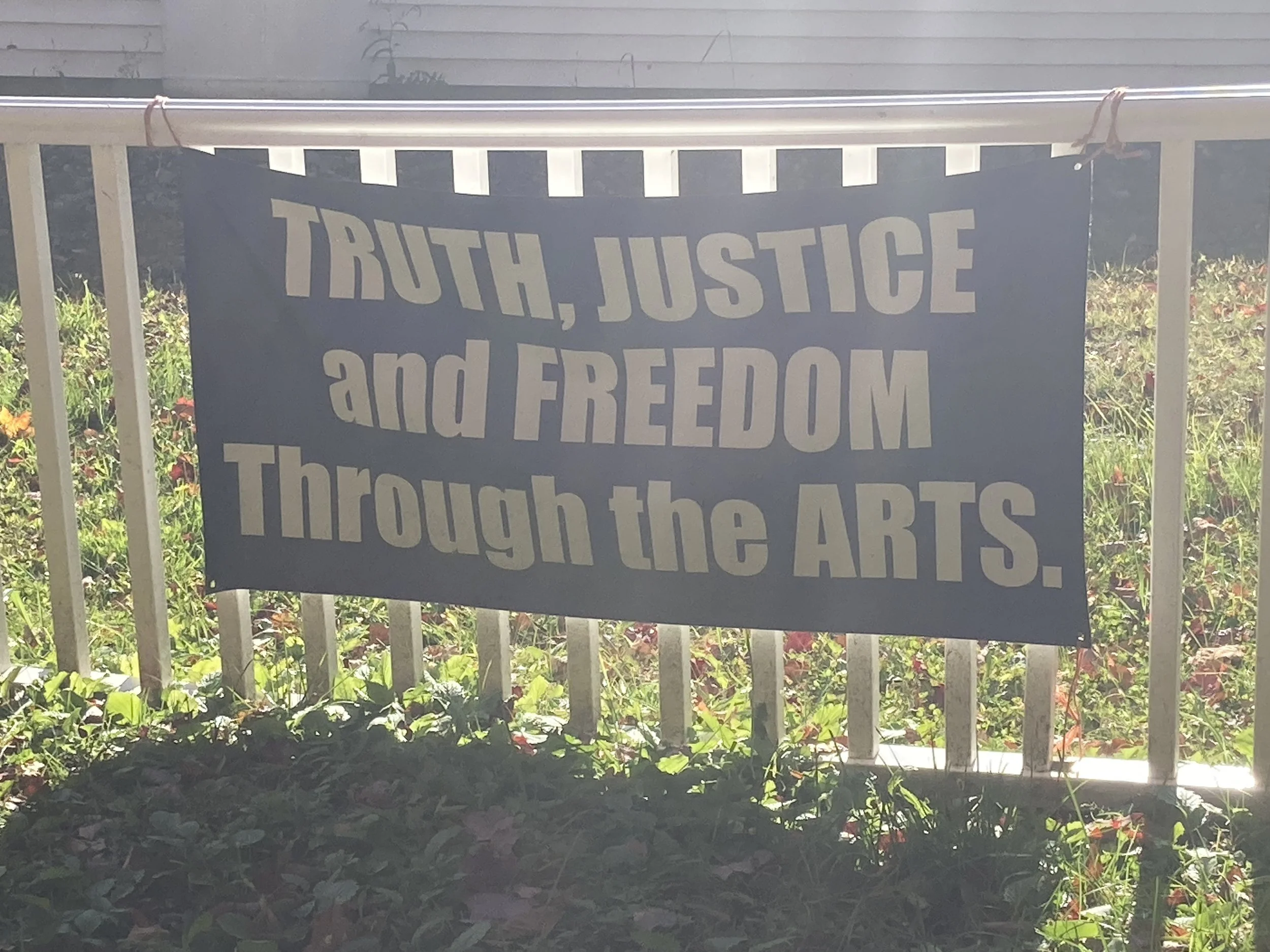 Dark blue sign reading “Truth, Justice and Freedom Through the Arts” displayed on a garden fence.
