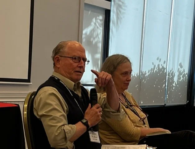 Two attorneys from the Coalition for Civil Freedoms. An older man speaking into a microphone and gesturing beside a seated woman at an indoor presentation.