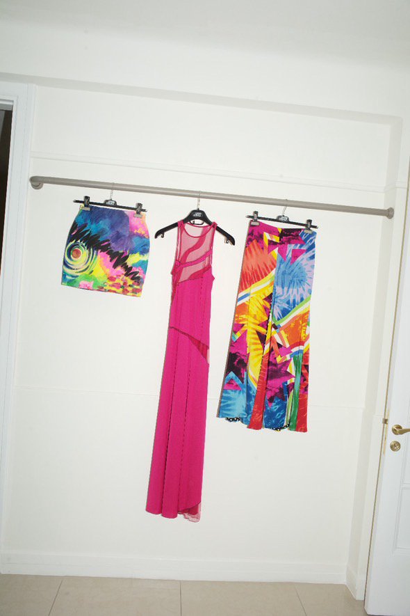 Colorful swimwear and a pink cover-up hanging on a silver clothing rod against a white wall.