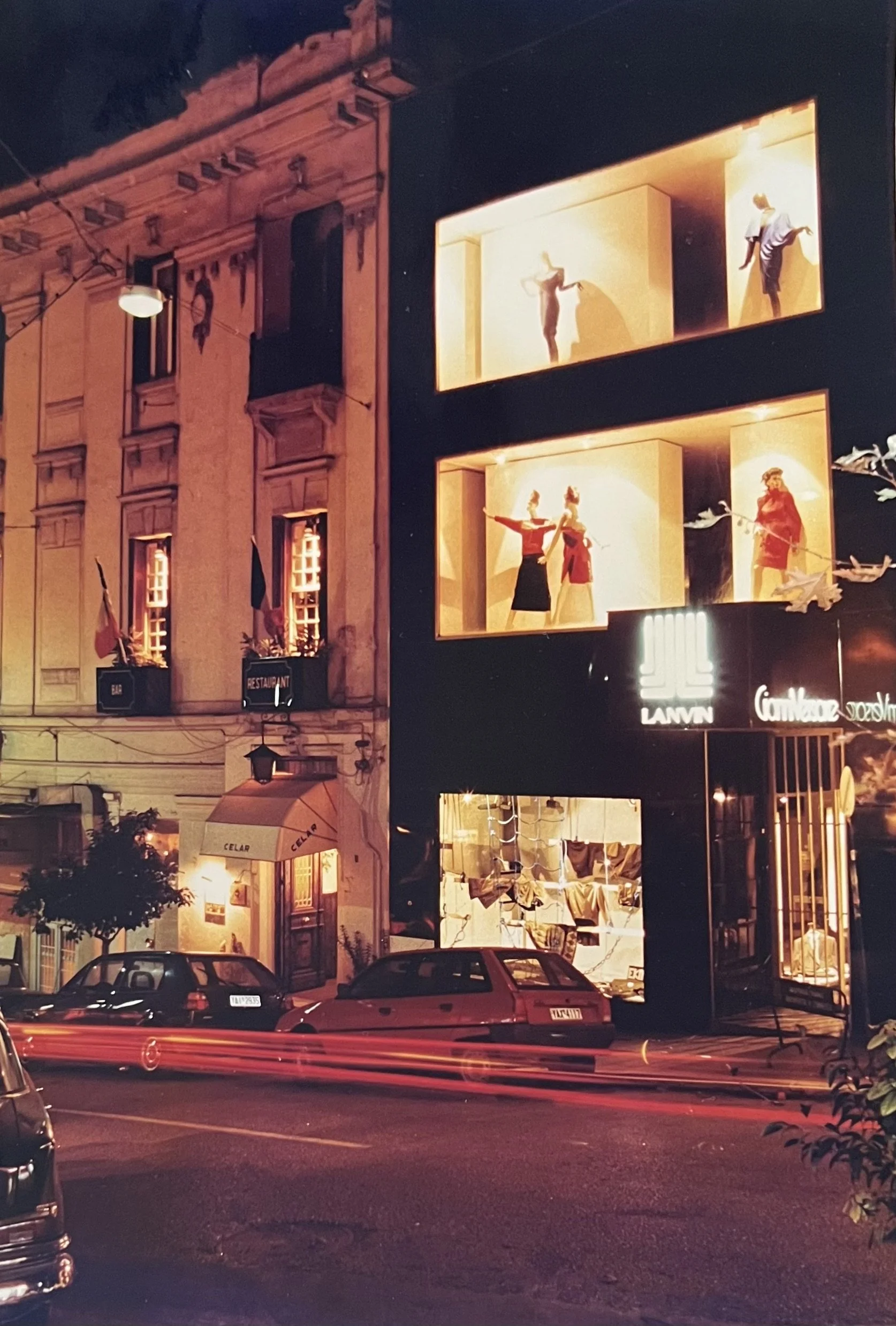 Nighttime street scene with parked cars, illuminated building with mannequins in display windows, and a restaurant sign that reads 'CELAR'