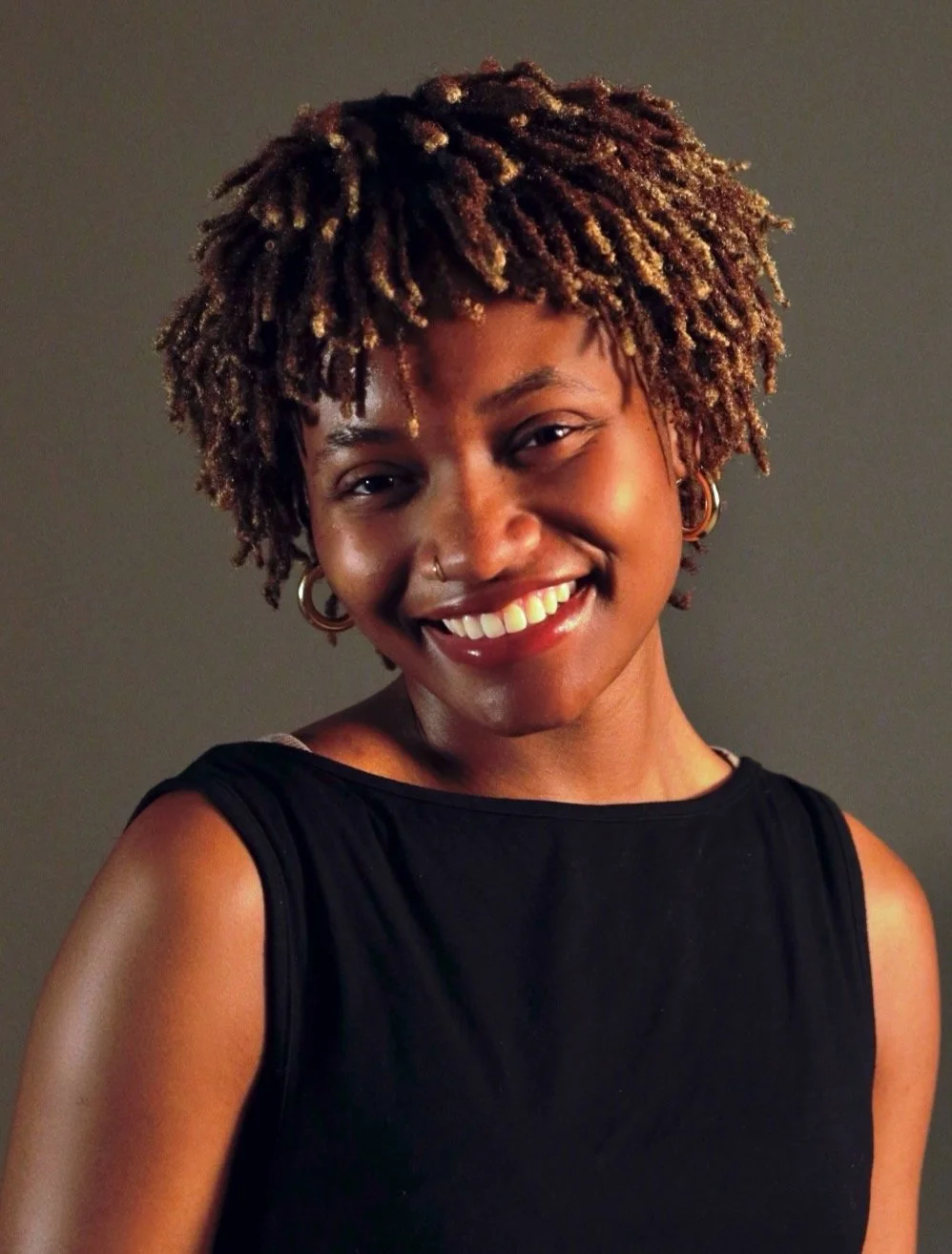 Smiling woman with short curly hair, large hoop earrings, and a nose ring, wearing a black sleeveless top.
