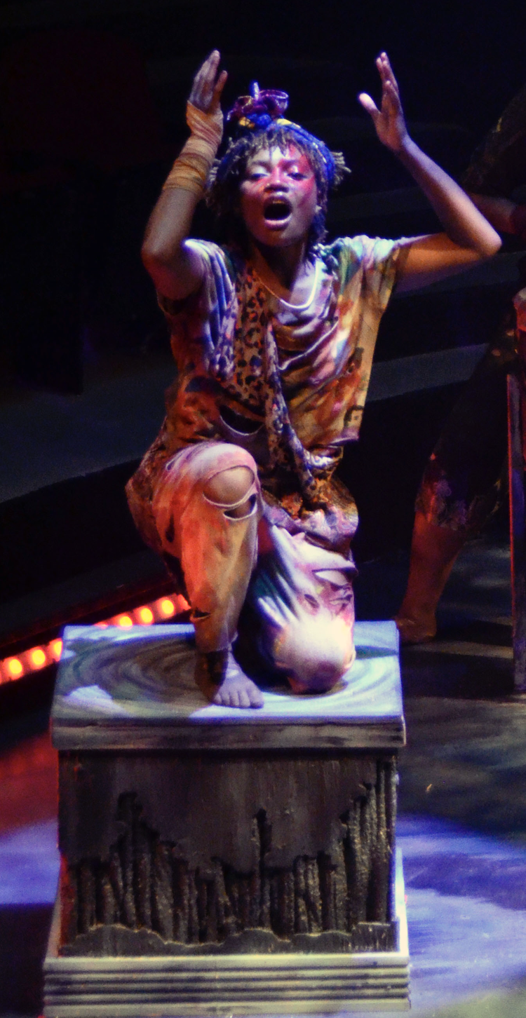 Performer with colorful clothing and dreadlocks kneeling on a prop that resembles a pedestal, with an expressive face and hands raised.