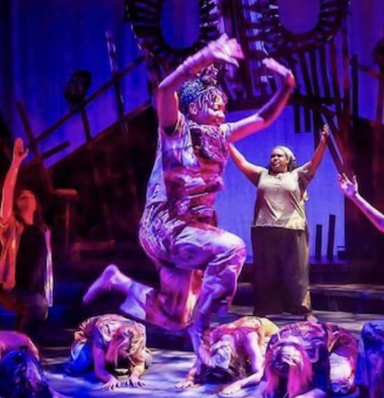 Performer balancing on one leg on stage with others kneeling and lying around, under colorful stage lighting.