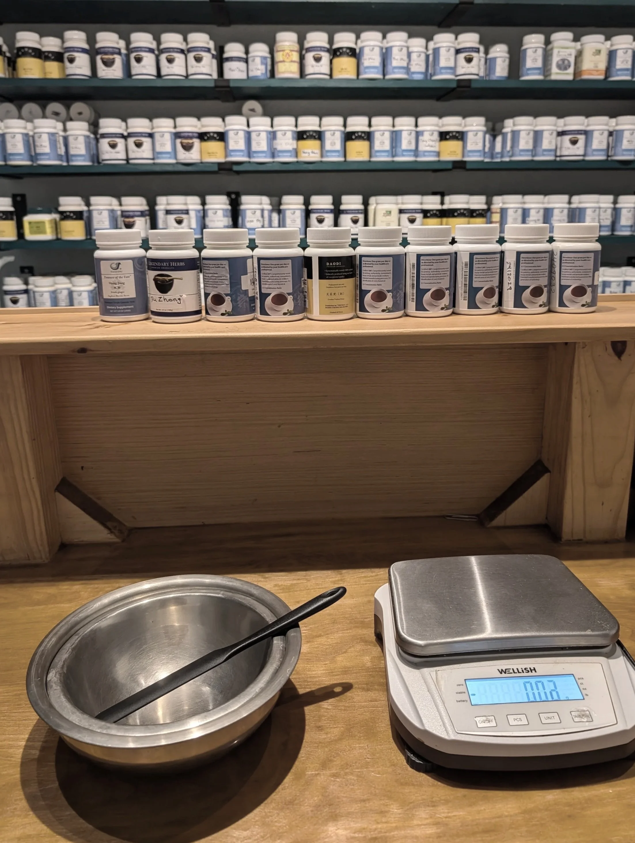 A kitchen or herbal shop scene with shelves filled with bottles of herbal supplements in the background, a wooden counter with a stainless steel bowl, a pair of tongs, and a digital scale displaying 0.02 grams.