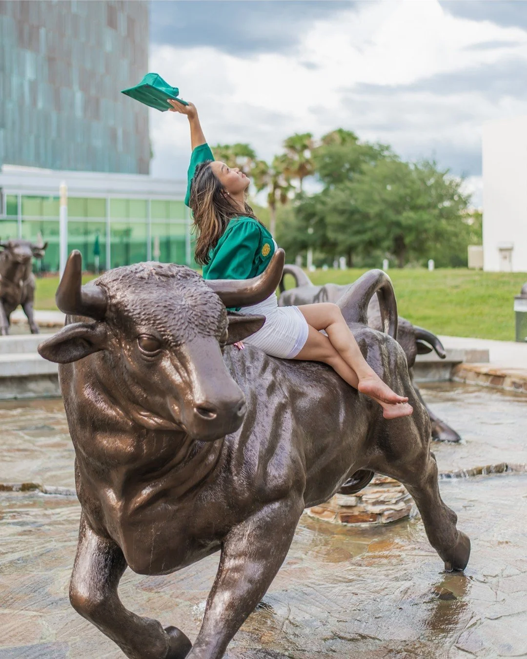 A woman sitting on a bronze bull sculpture in a fountain, reaching up with one arm holding a green object, outdoors in a park with trees and a cloudy sky in the background.