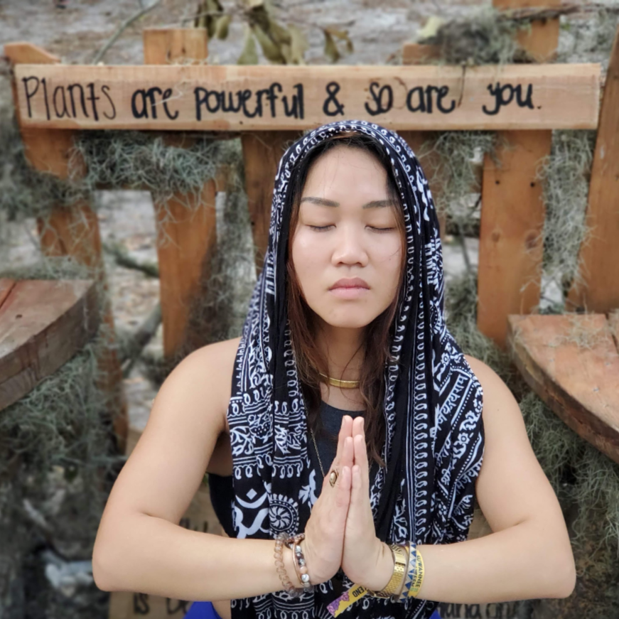 A woman with long brown hair, wearing a black and white patterned headscarf, is meditating with her eyes closed and hands in a prayer position. Behind her is a wooden sign that reads 'Plants are powerful & so are you,' in a natural outdoor setting with trees and moss.