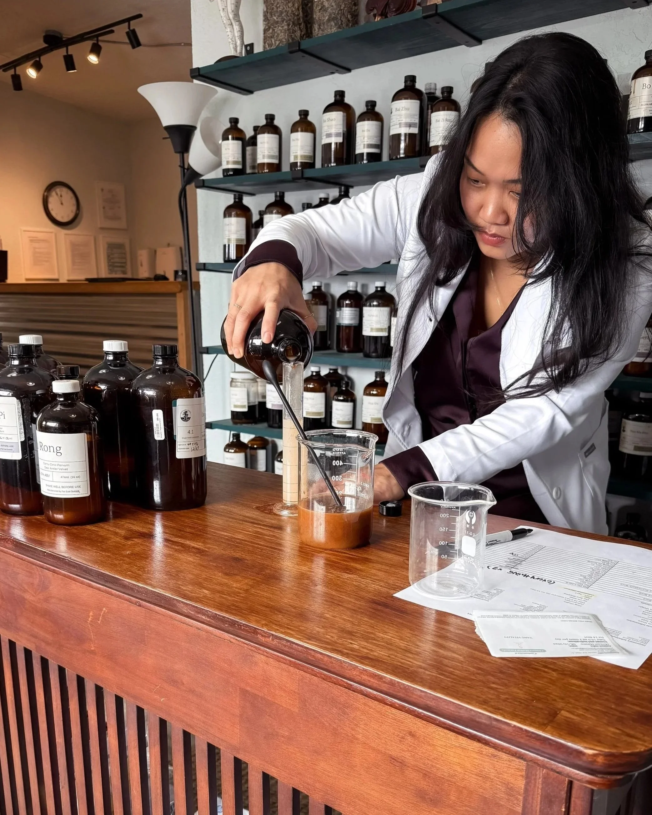 A woman in a white lab coat is working in a laboratory or apothecary, pouring a liquid from a small brown glass bottle into a beaker on a wooden counter. Behind her are shelves filled with similar brown bottles labeled with various names. On the counter, there are additional empty beakers, a pen, and a paper with handwritten notes. The environment appears organized and professional, with a warm lighting ambiance and a clock on the wall in the background.