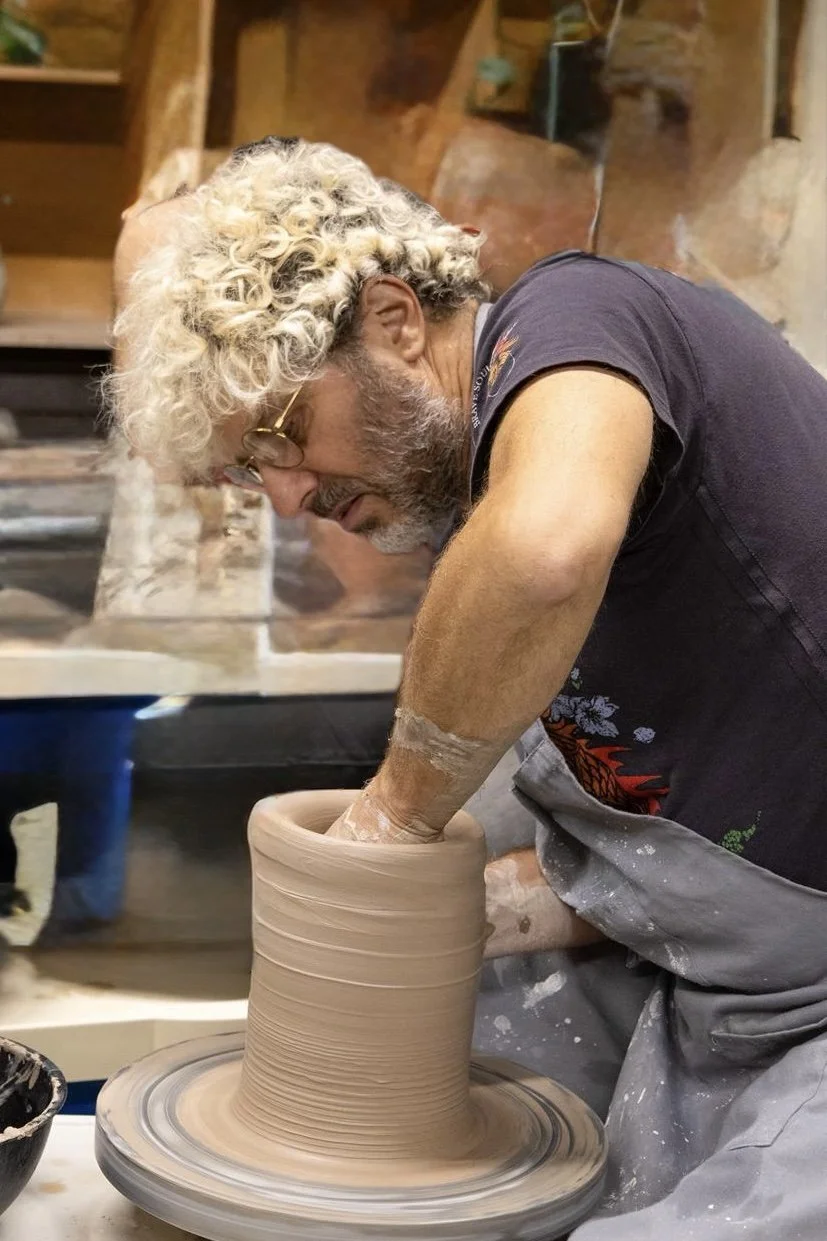 A man with curly blond hair and glasses shaping a ceramic pot on a pottery wheel in a workshop.