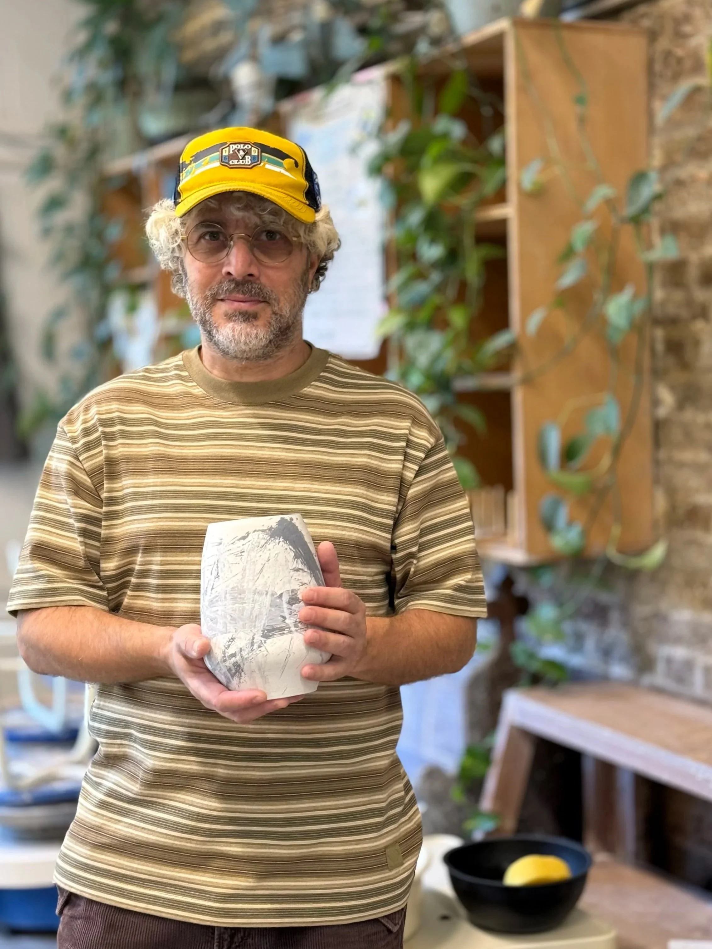A man with gray curly hair, beard, and glasses wearing a yellow cap and striped beige shirt holds a ceramic bowl in a pottery studio. The studio has shelves, green plants, and a brick wall in the background.