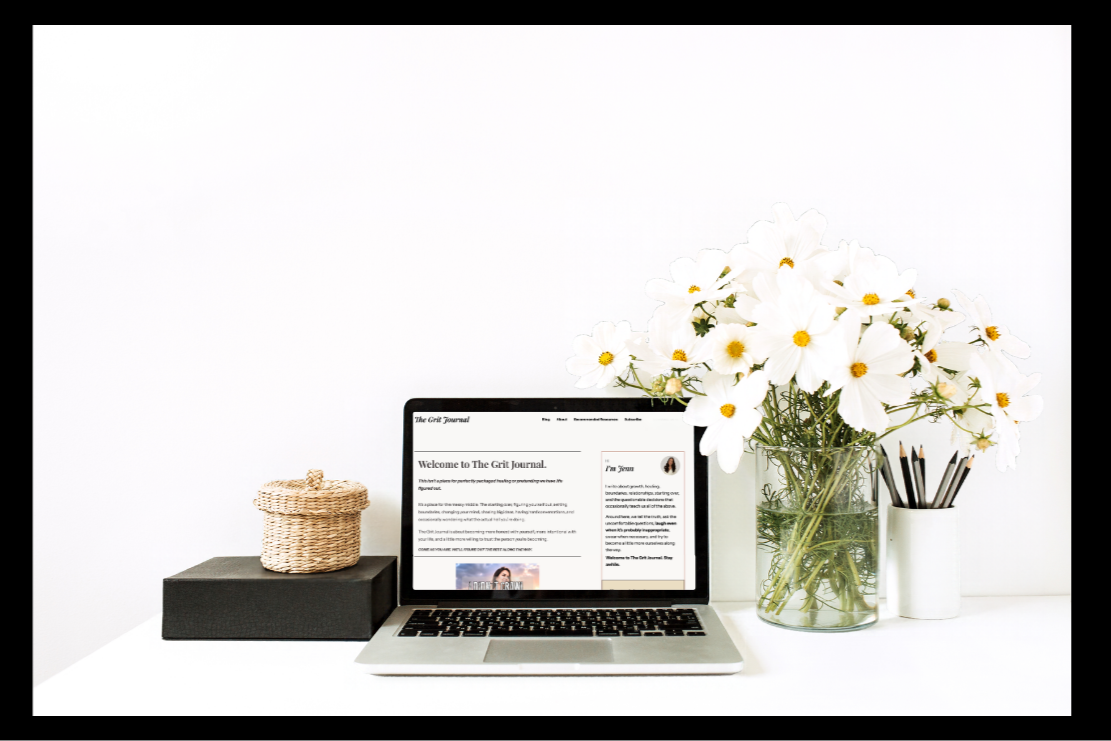 The Grit Journal website by Jenn Armstrong displayed on a laptop, surrounded by white flowers and minimalist desk accessories.