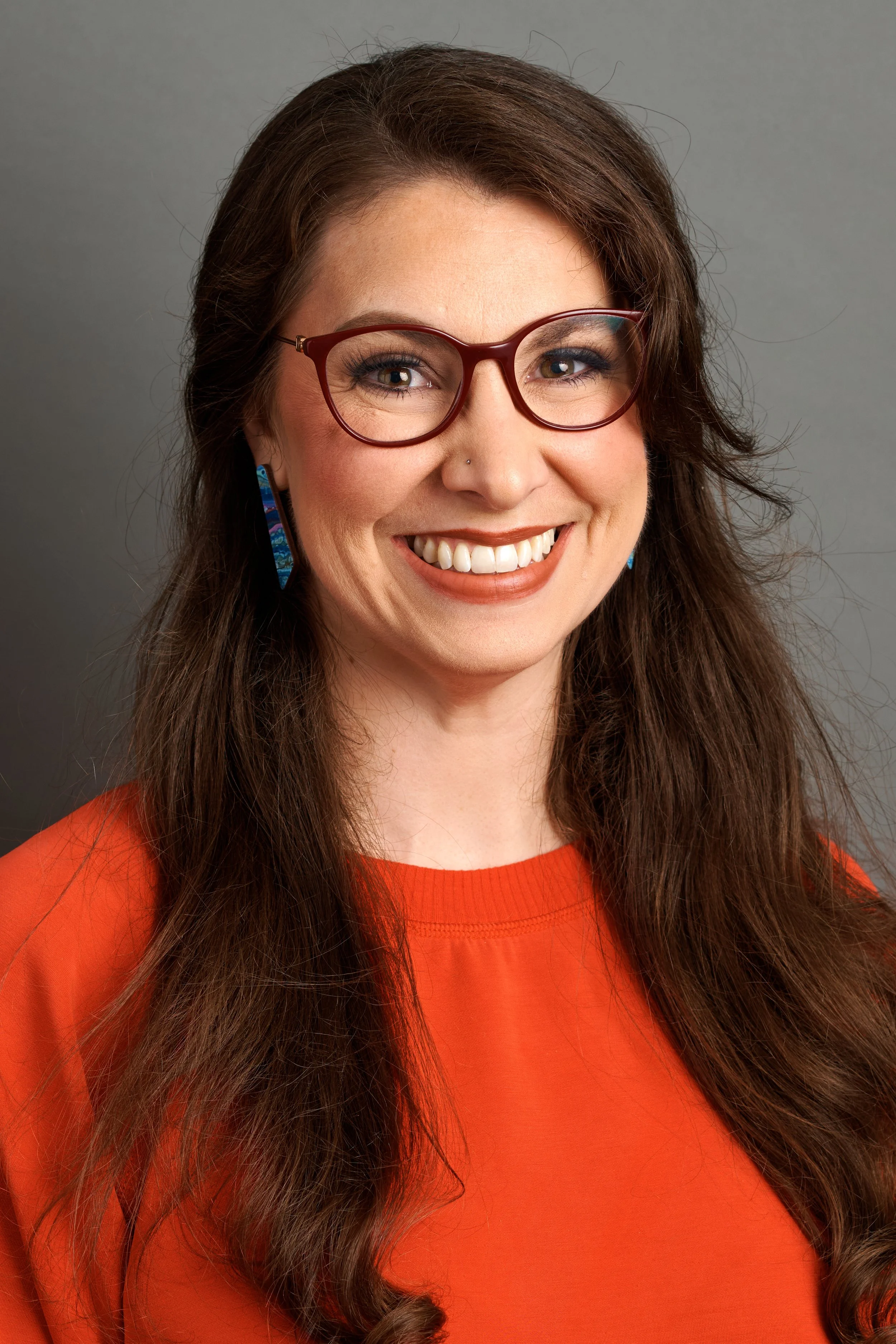 A woman with long, wavy brown hair wearing glasses with a red frame, earrings, and a nose piercing, smiling against a plain gray background, dressed in an orange top.