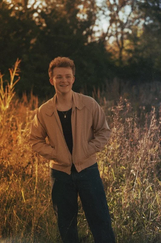 A young man with short, curly hair smiling and wearing a beige jacket over a black shirt, standing in a field with tall grass and trees in the background during sunset.