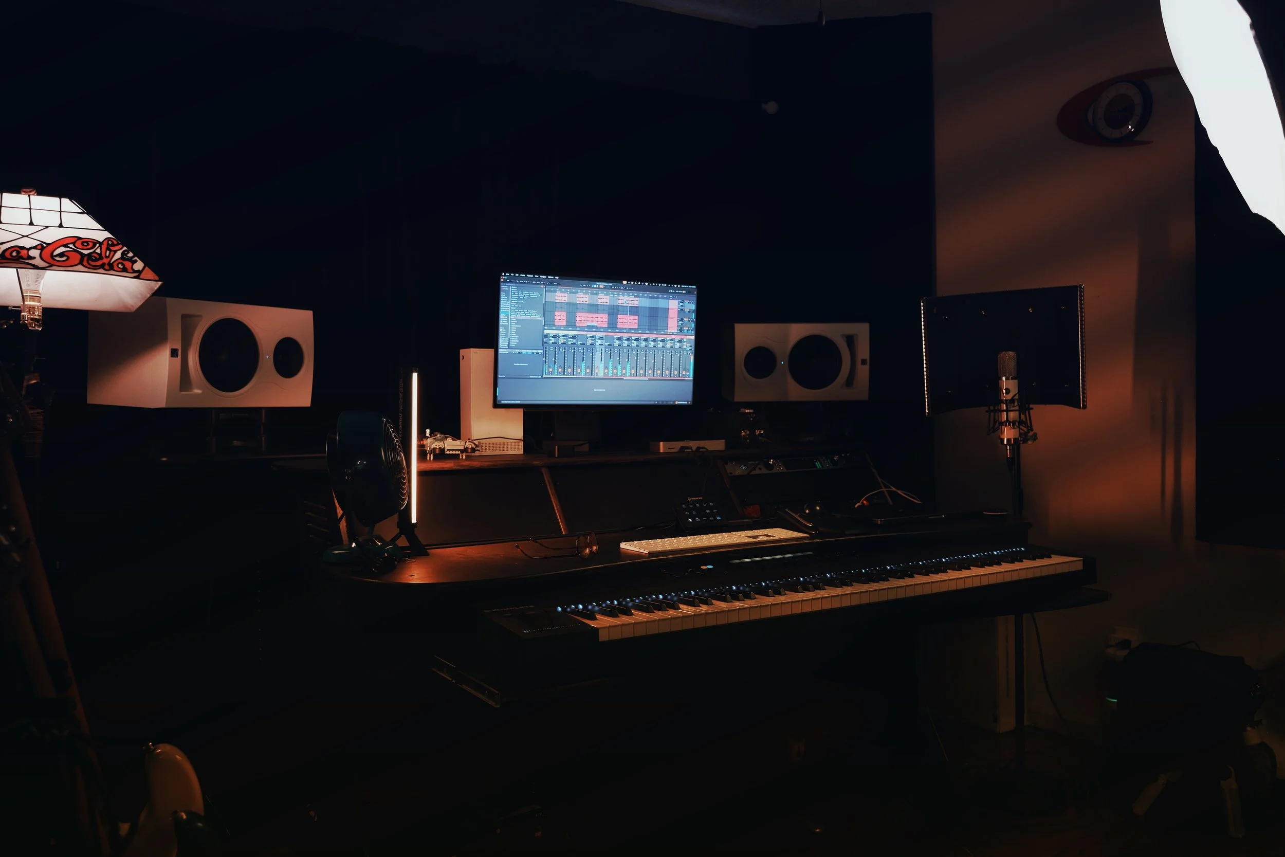Darkly lit music production studio with a computer monitor displaying digital audio workstation software, studio speakers, a keyboard, and various audio equipment and microphones.