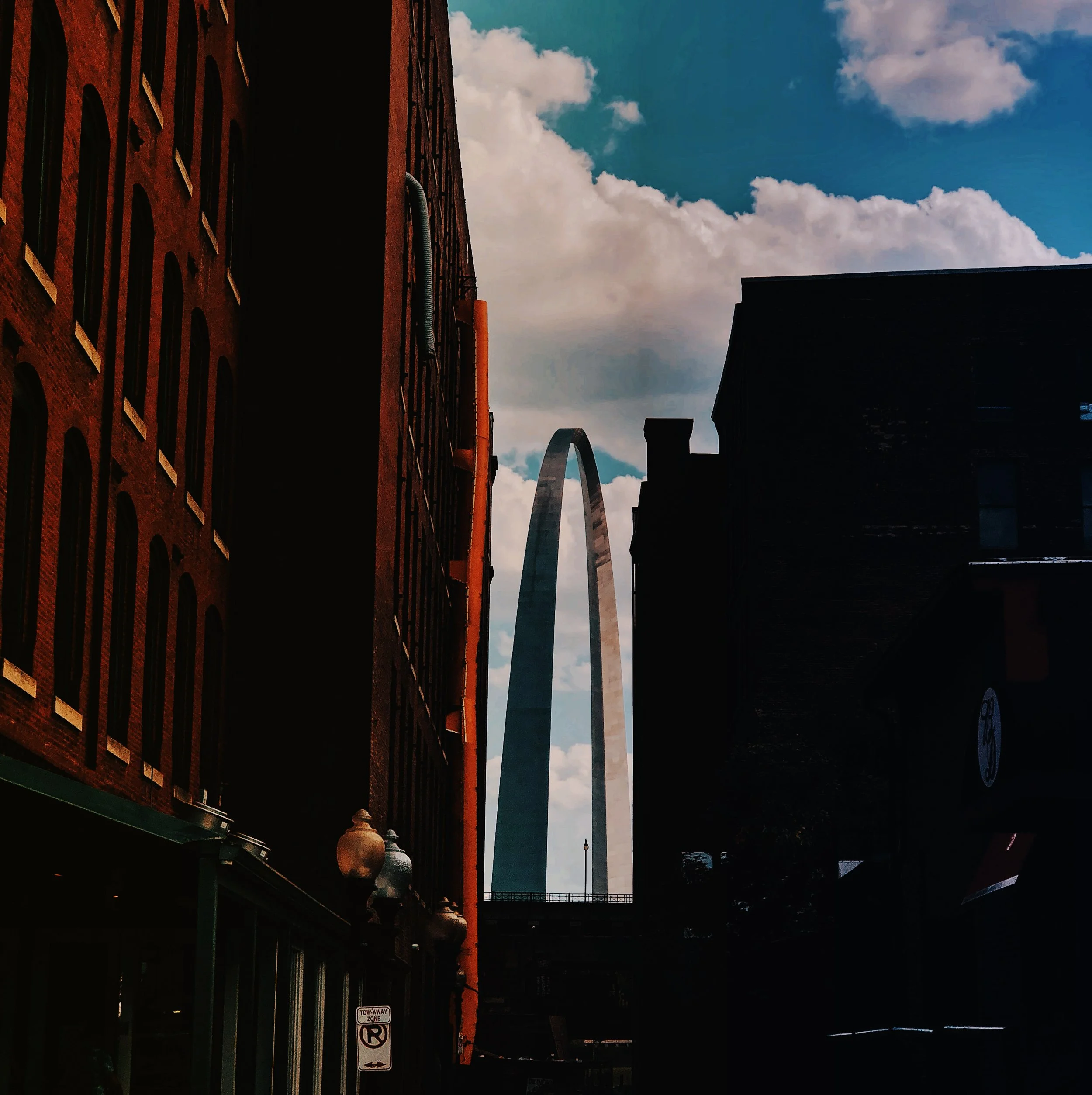 Street scene at sunset with red brick buildings on the left, a street sign prohibiting parking, and the Gateway Arch in the background against a partly cloudy sky.