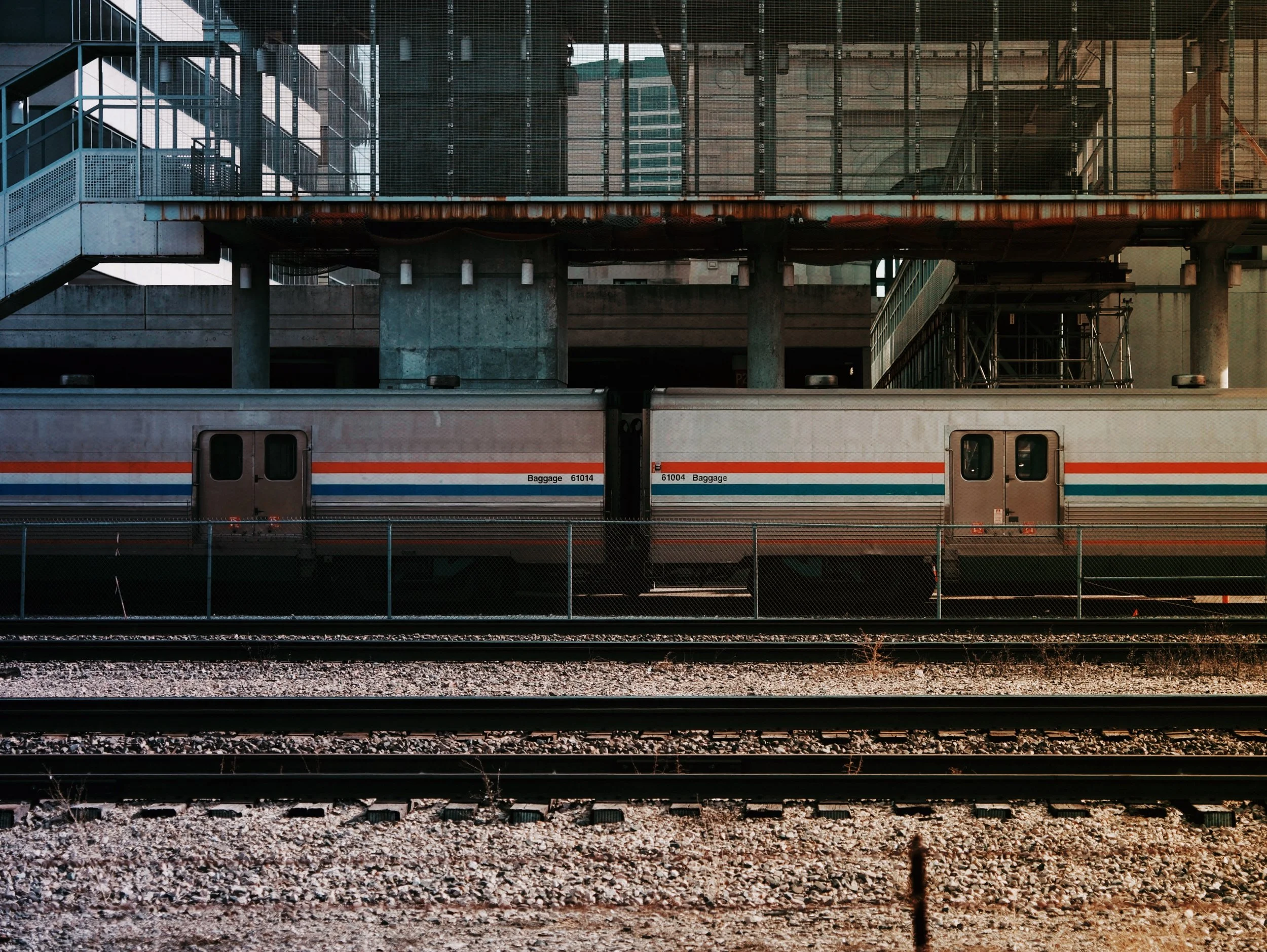 View of a train station platform with a passing train. The train has a gray exterior with red and blue stripes and the word 'Baggage' and numbers on it. Above the platform, there is an industrial-style overpass with metal stairs, railings, and scaffo